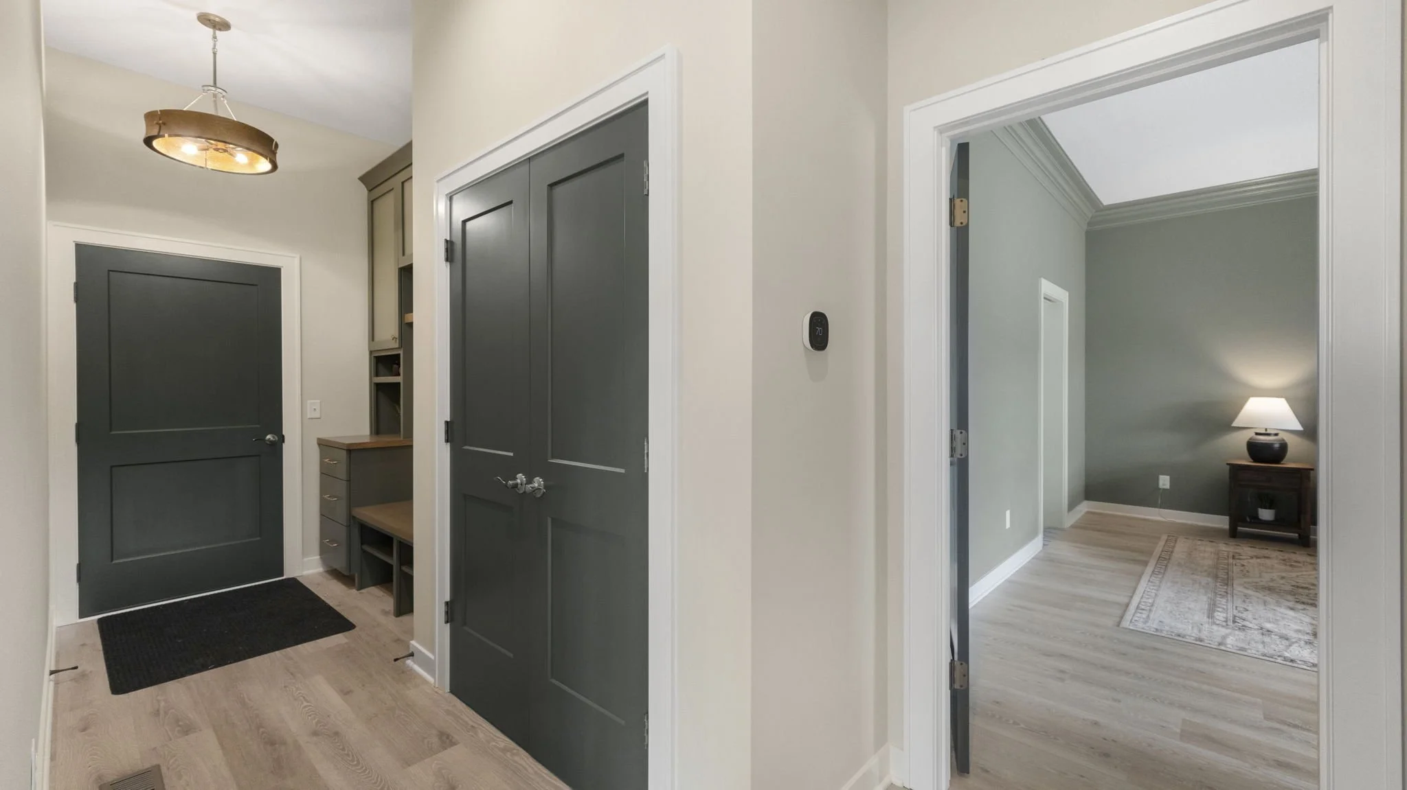 Entryway with dark gray double doors, a small wooden cabinet with drawers and open shelves, a black doormat, and a hallway leading to a living room with a green wall, a rug, a side table, and a lamp.