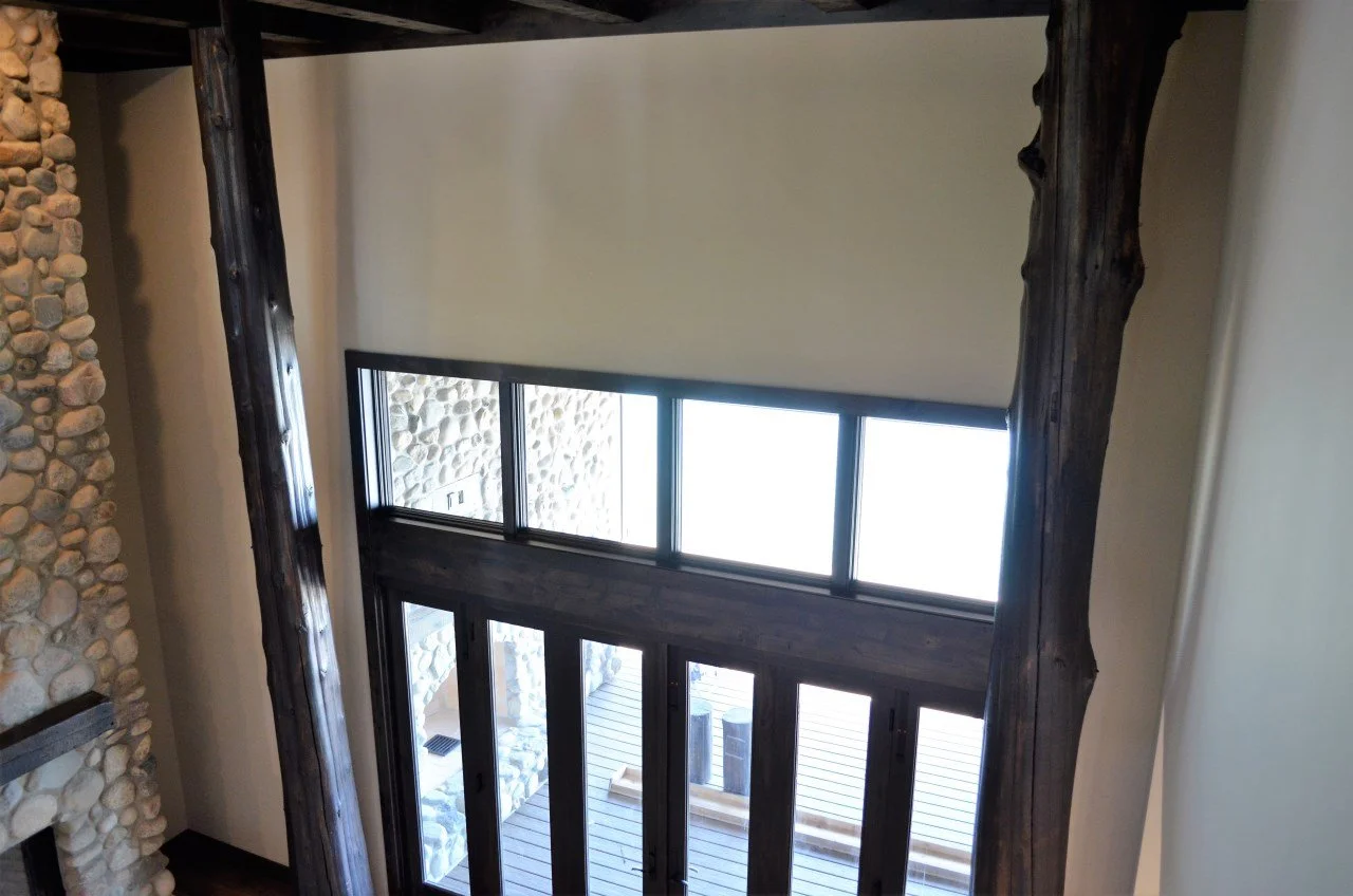 Interior view of a room showing large windows and a wooden beam structure, with a stone fireplace on the left and a deck outside.