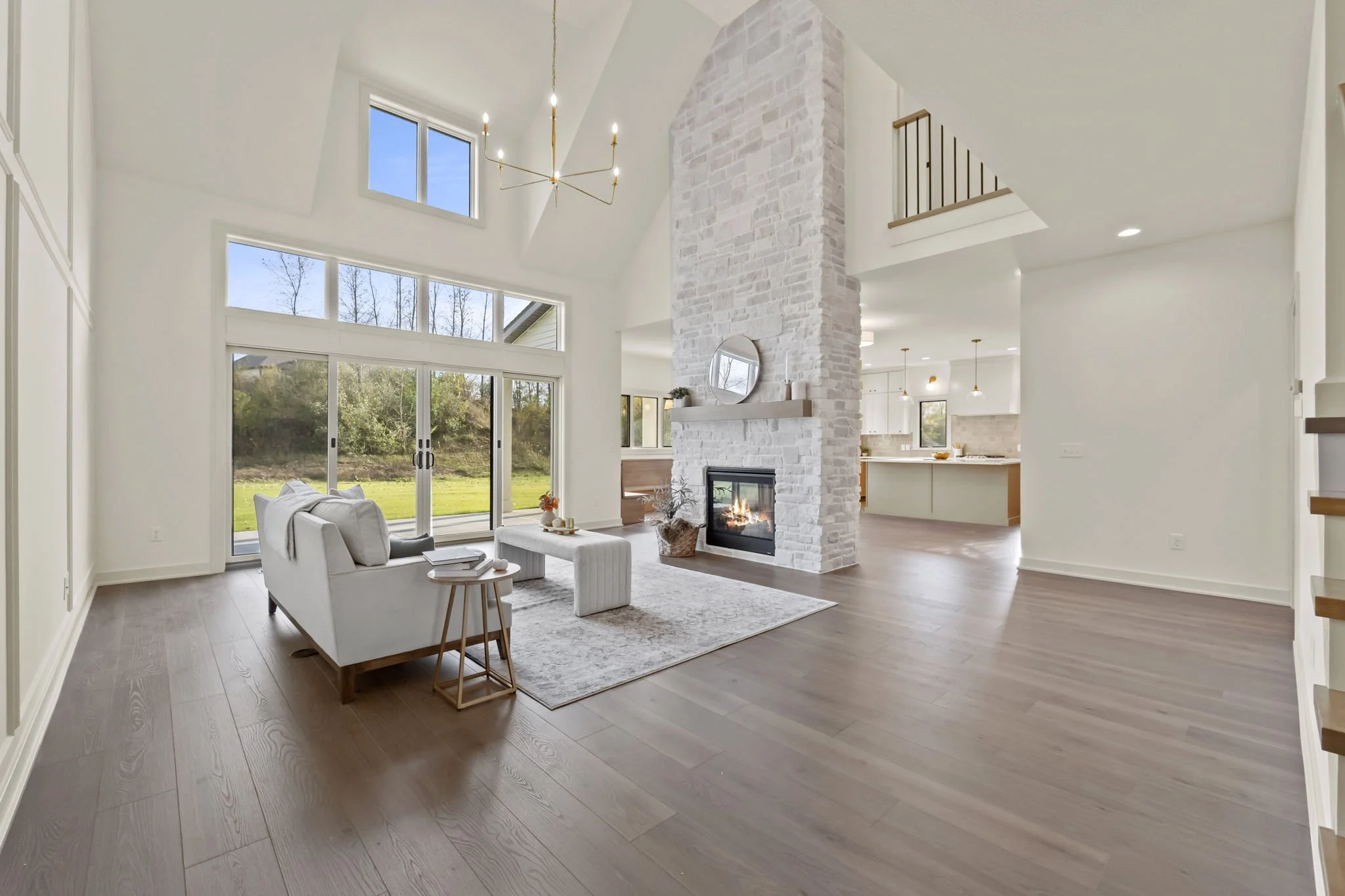 Bright living room with high ceilings, white walls, and large windows overlooking a grassy backyard. A contemporary fireplace is built into a white brick chimney with a circular mirror above. The room features a white sofa, a small wooden side table,