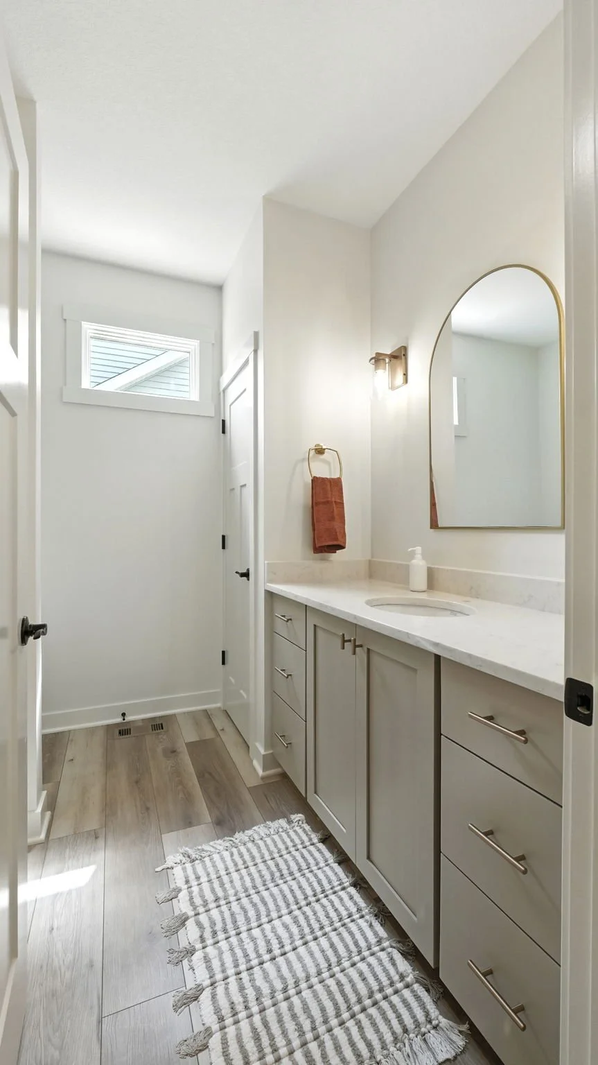 A modern bathroom with light-colored cabinets, a white countertop, and a large mirror with a gold frame. There is a reddish-brown towel hanging on a gold towel ring, and a striped rug on the wooden floor. A small window is visible on the upper left s