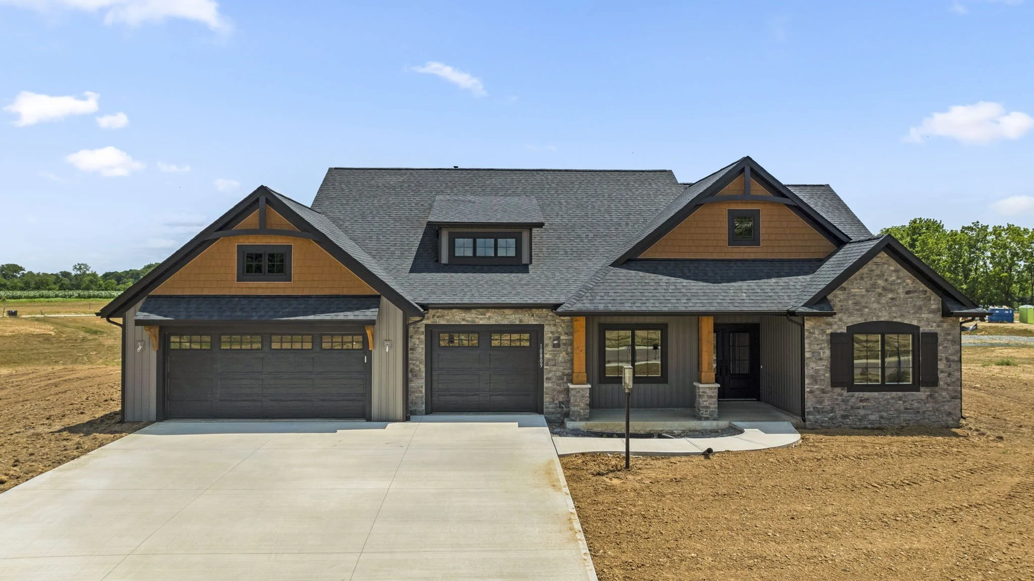 Newly built house with a multi-garage on a concrete driveway, a front porch with columns, and a mixed exterior of wood, stone, and siding. The house is in a rural area with fields and trees in the background under a clear blue sky.