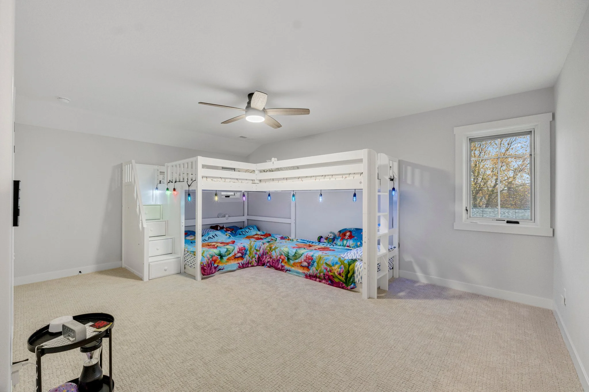 A children's bedroom with a white bunk bed decorated with colorful ocean-themed bedding, string lights, a single window showing trees outside, and a beige carpeted floor.