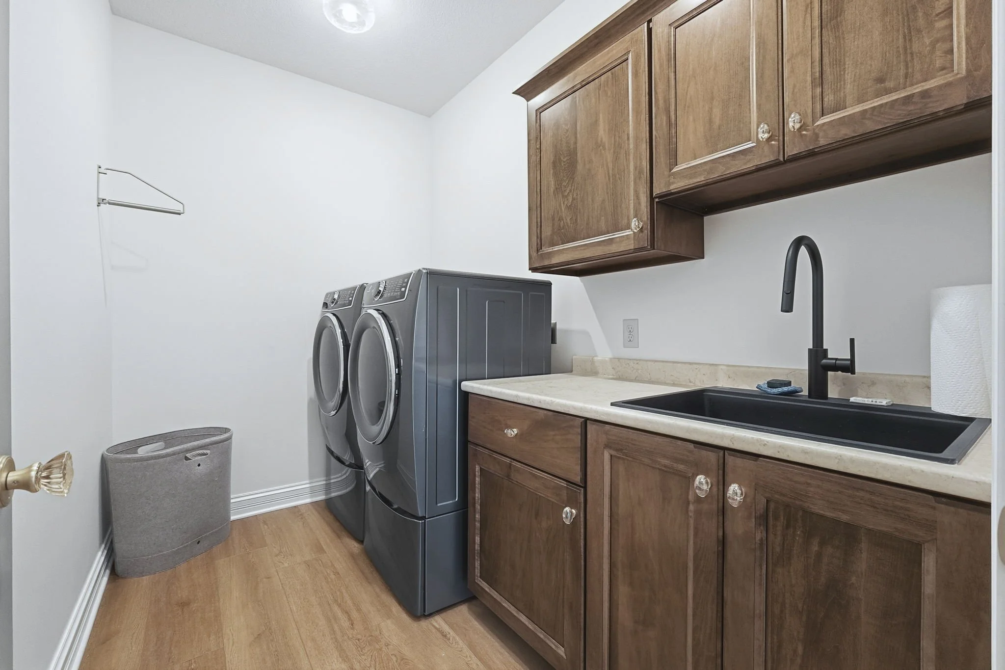 Laundry room with a washing machine, a dryer, wooden cabinets, a black sink, and a paper towel roll.