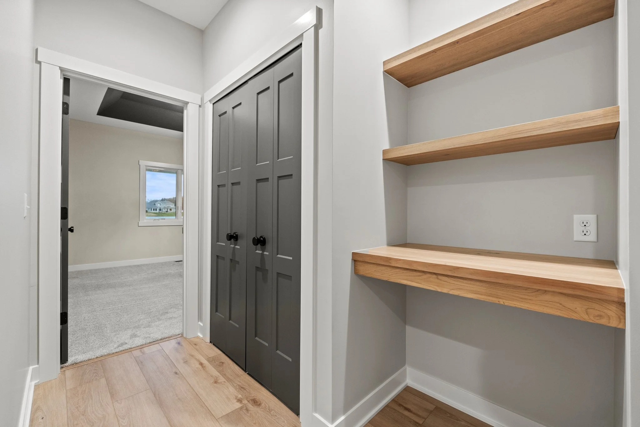 A hallway view showing a closet with dark gray double doors, a doorway leading to a room with a window, light wood flooring, and a wall with three wooden shelves and an electrical outlet.