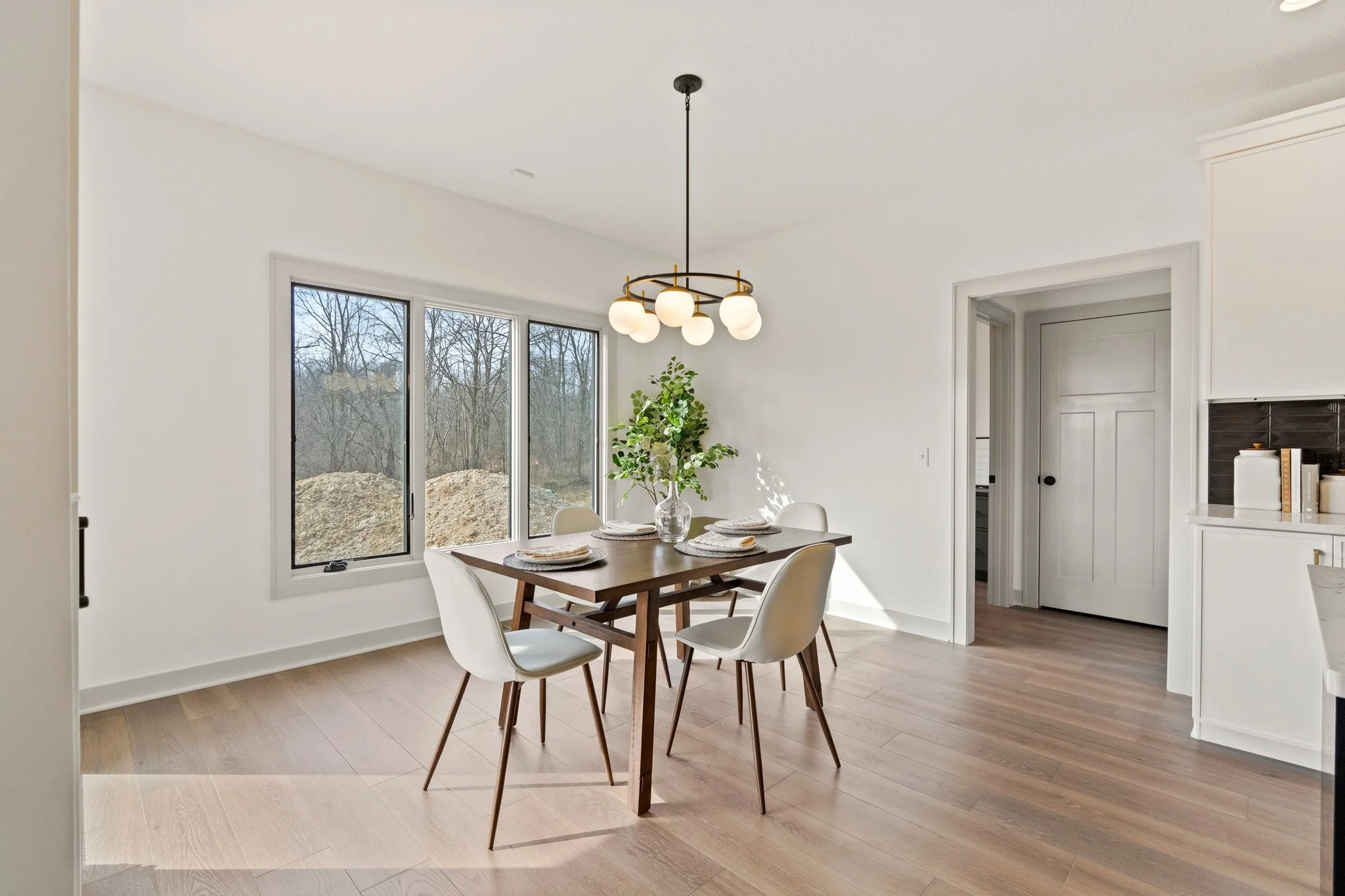 Dining room with a wooden table, four white chairs, a centerpiece with greenery, large window showing an outdoor landscape, and modern light fixture