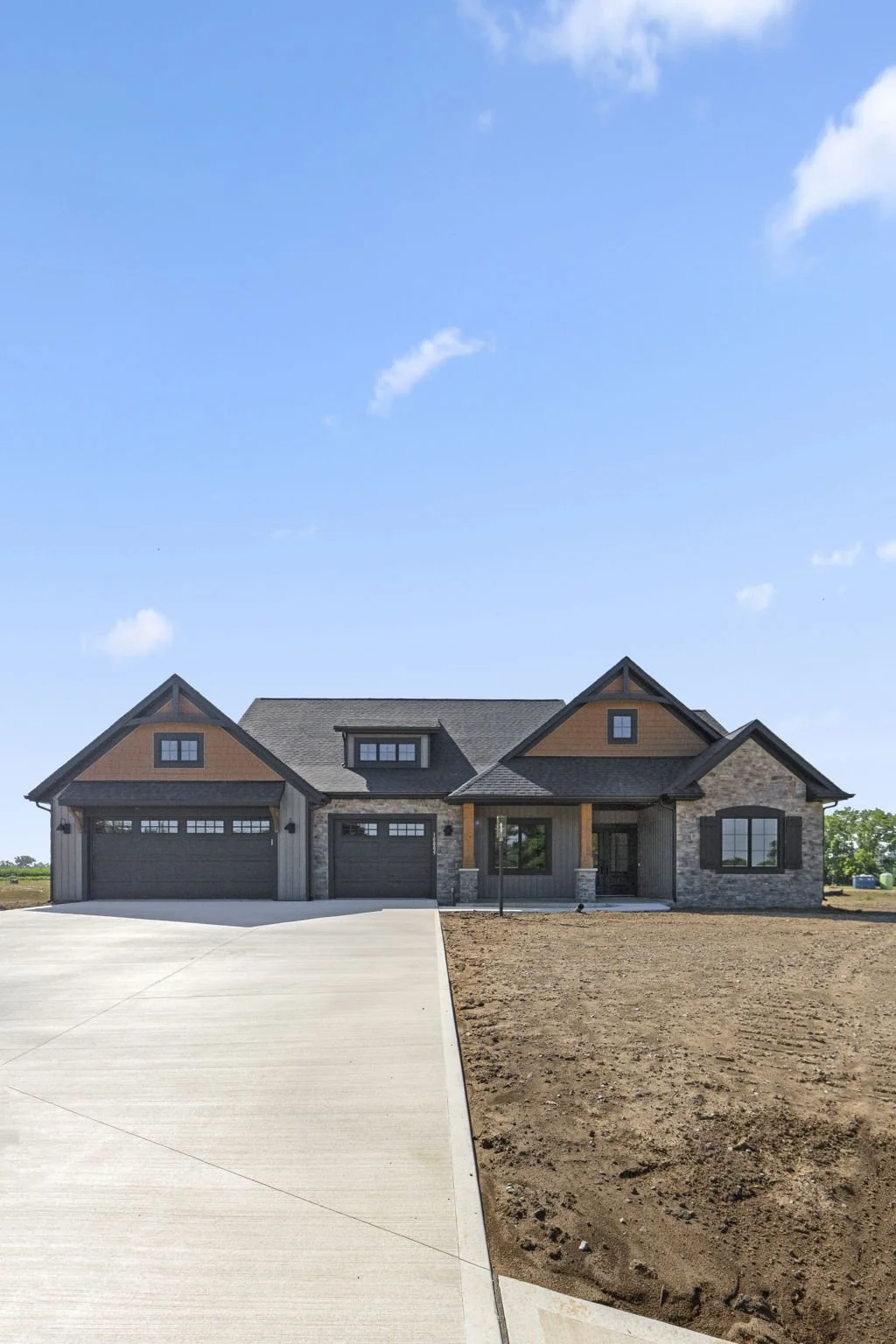New house under construction with a large concrete driveway, stone and wood exterior, and a clear blue sky.