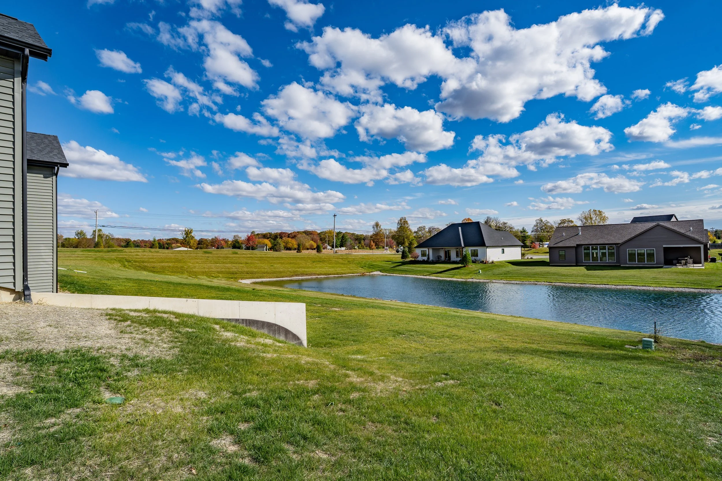 A suburban neighborhood with a pond in the middle, surrounded by houses and green lawns under a blue sky with scattered clouds.