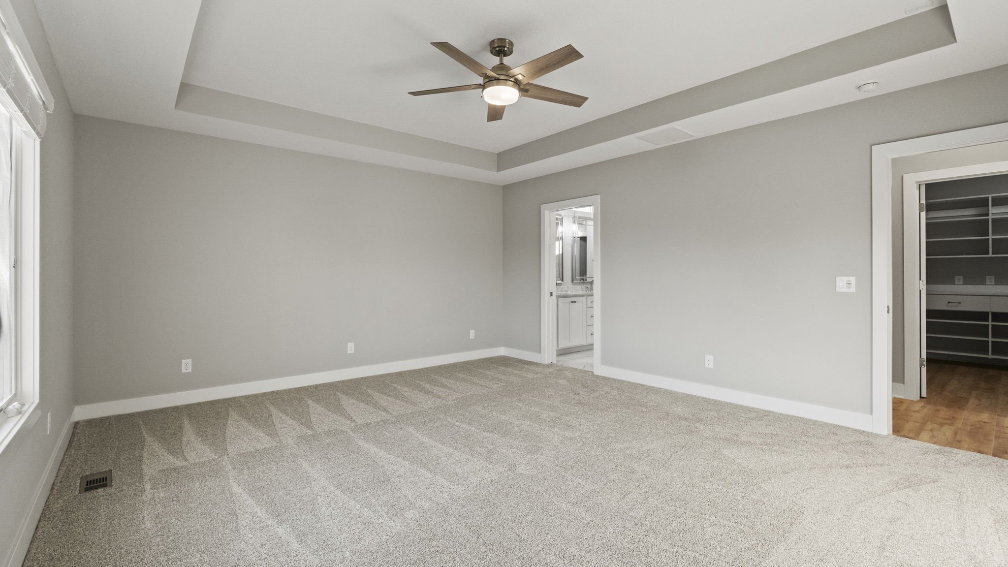 Empty room in a house with light gray walls, beige carpet, a ceiling fan, and two doorways, one leading to a bathroom and another to a walk-in closet.