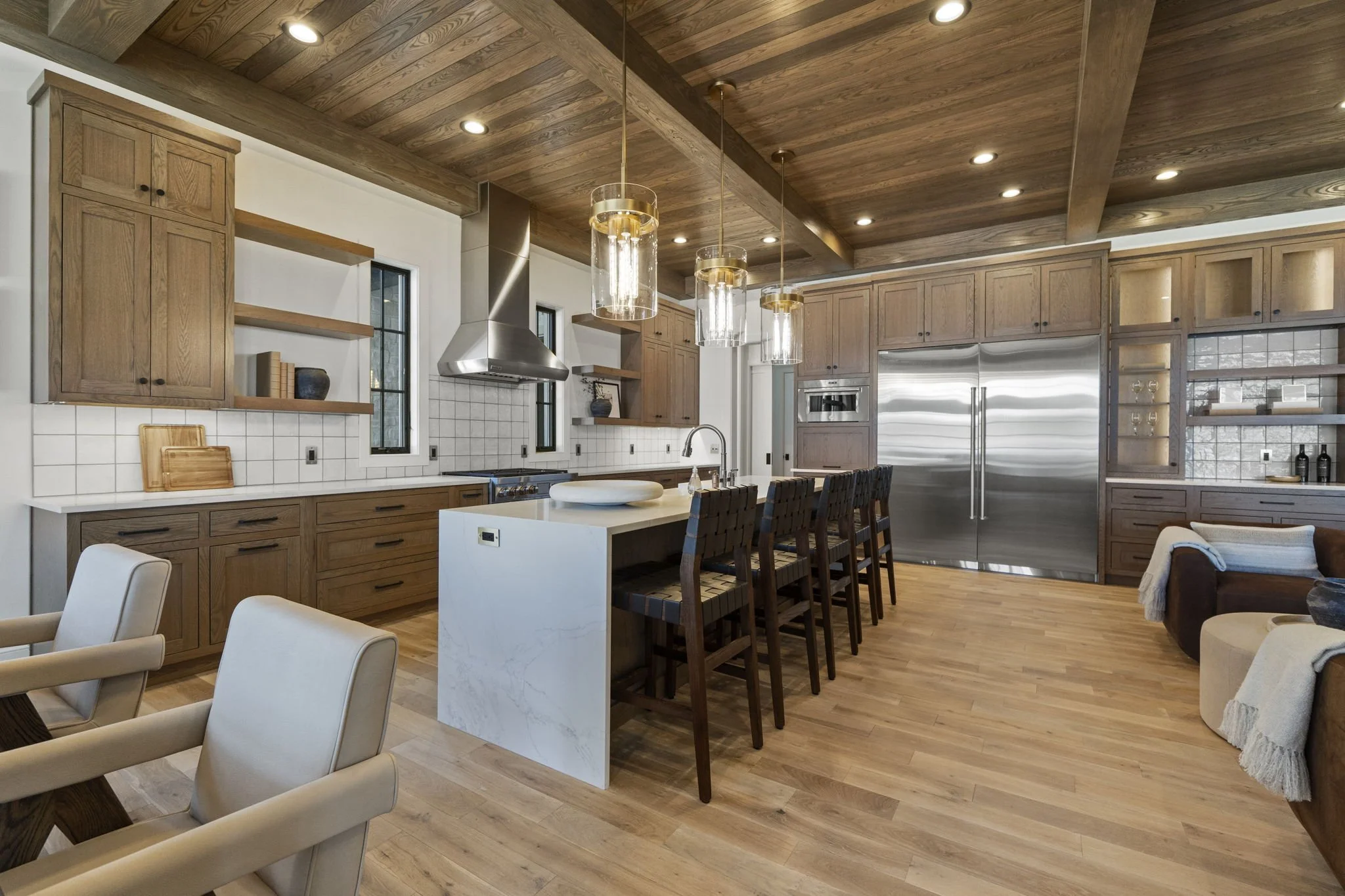Modern kitchen with wooden cabinets, white tile backsplash, stainless steel appliances, and an island with bar seating, featuring a wood-paneled ceiling and hardwood floor.