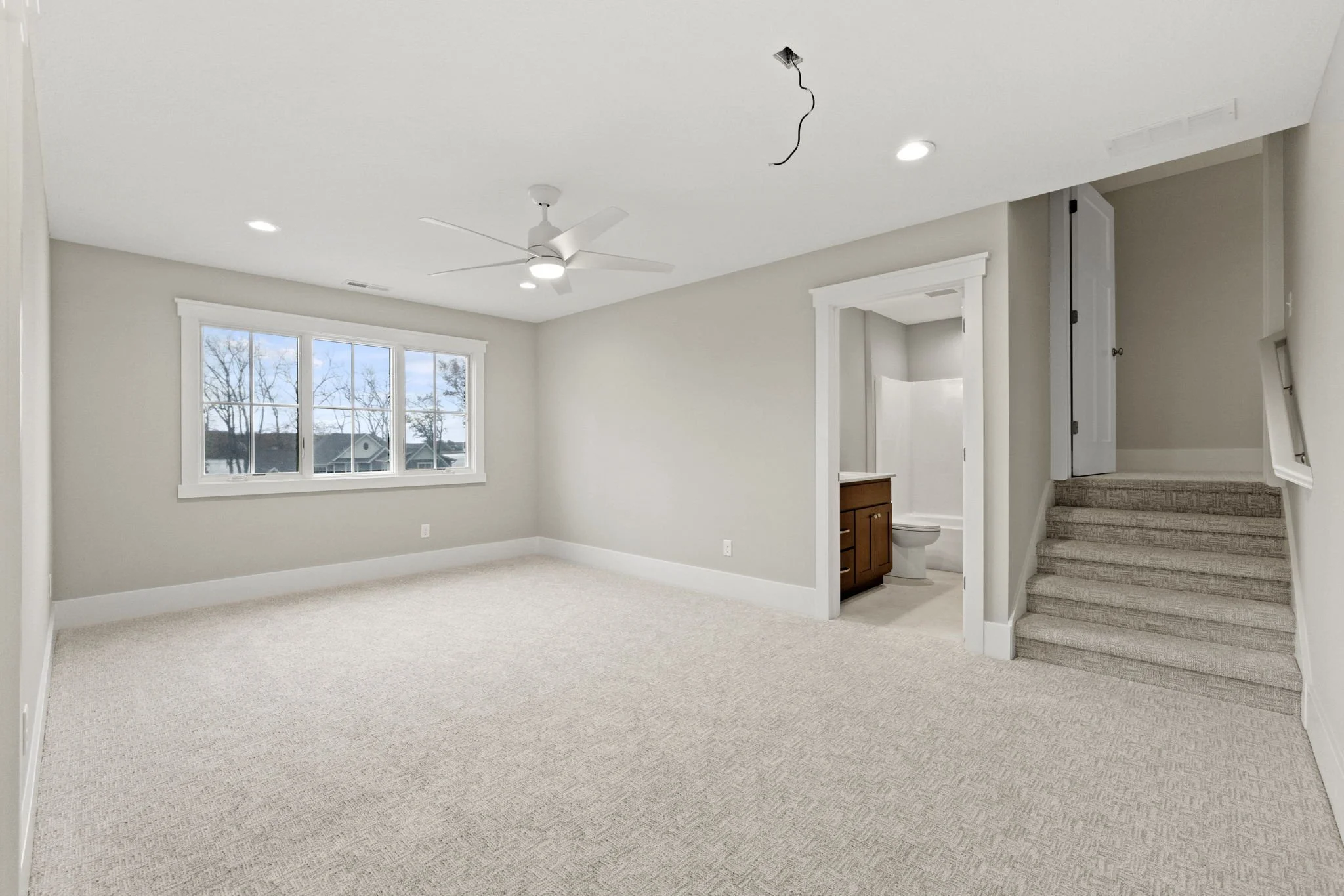 Empty room with beige carpet, white walls, large window, ceiling fan, and an adjacent bathroom with wooden vanity and toilet. Carpeted stairs and a small closet near the stairs.