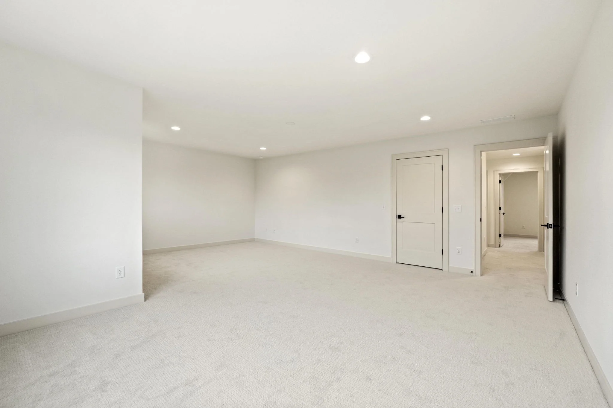 Empty room with white walls, ceiling, and carpeted floor, with closed white door and open doorway leading to another room, lit by ceiling lights.
