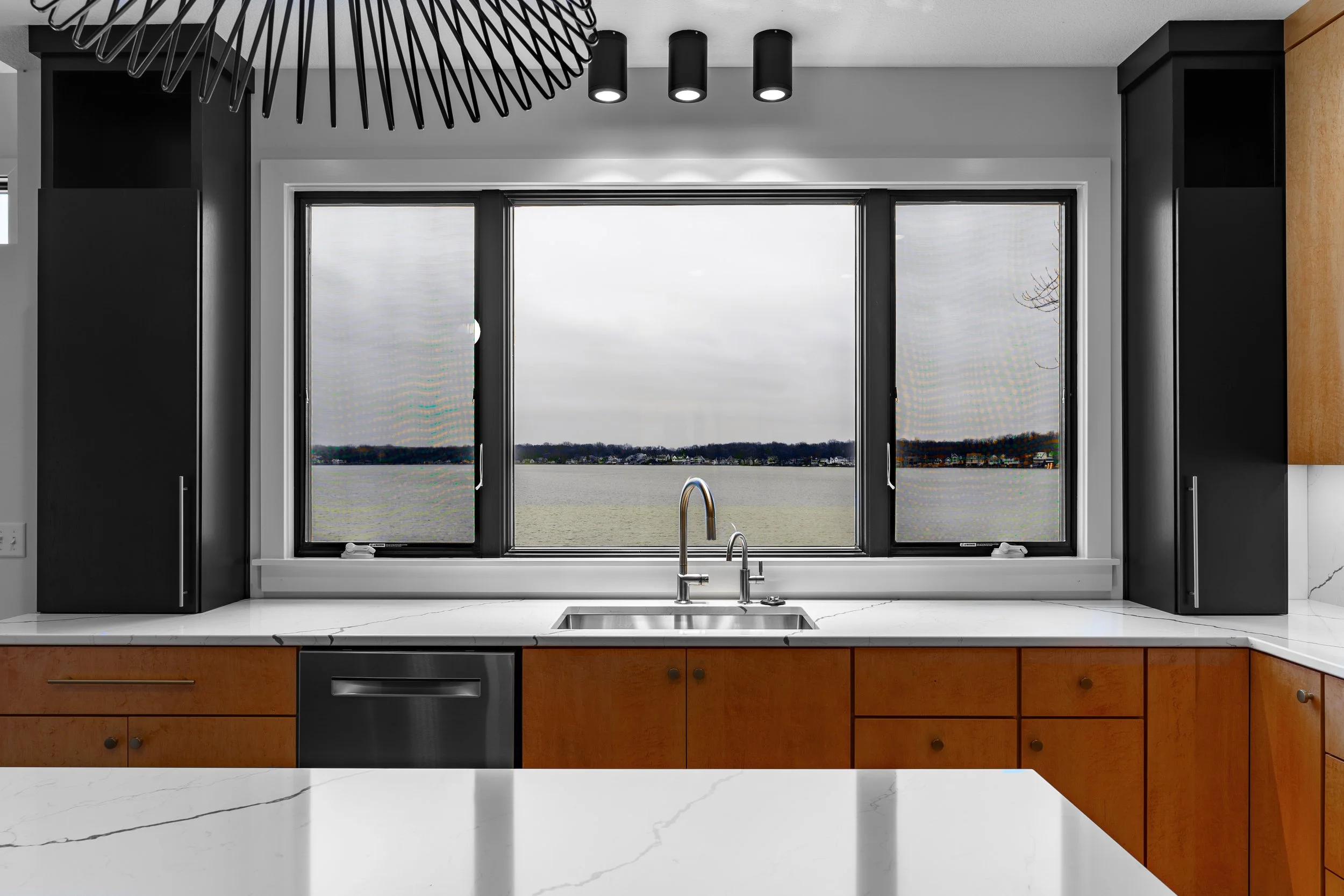 Modern kitchen with white marble countertops, wooden cabinets, black upper cabinets, and a large window overlooking a lake.
