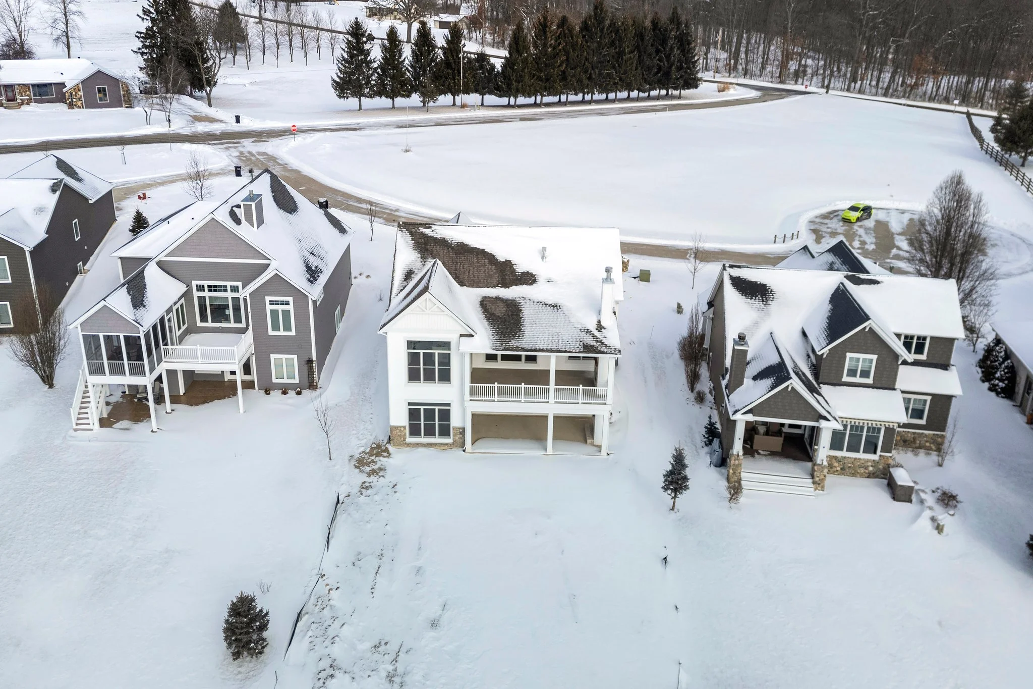 An aerial view of three houses in a snowy neighborhood, with a snow-covered field and trees in the background, and a car parked on the driveway of one house.