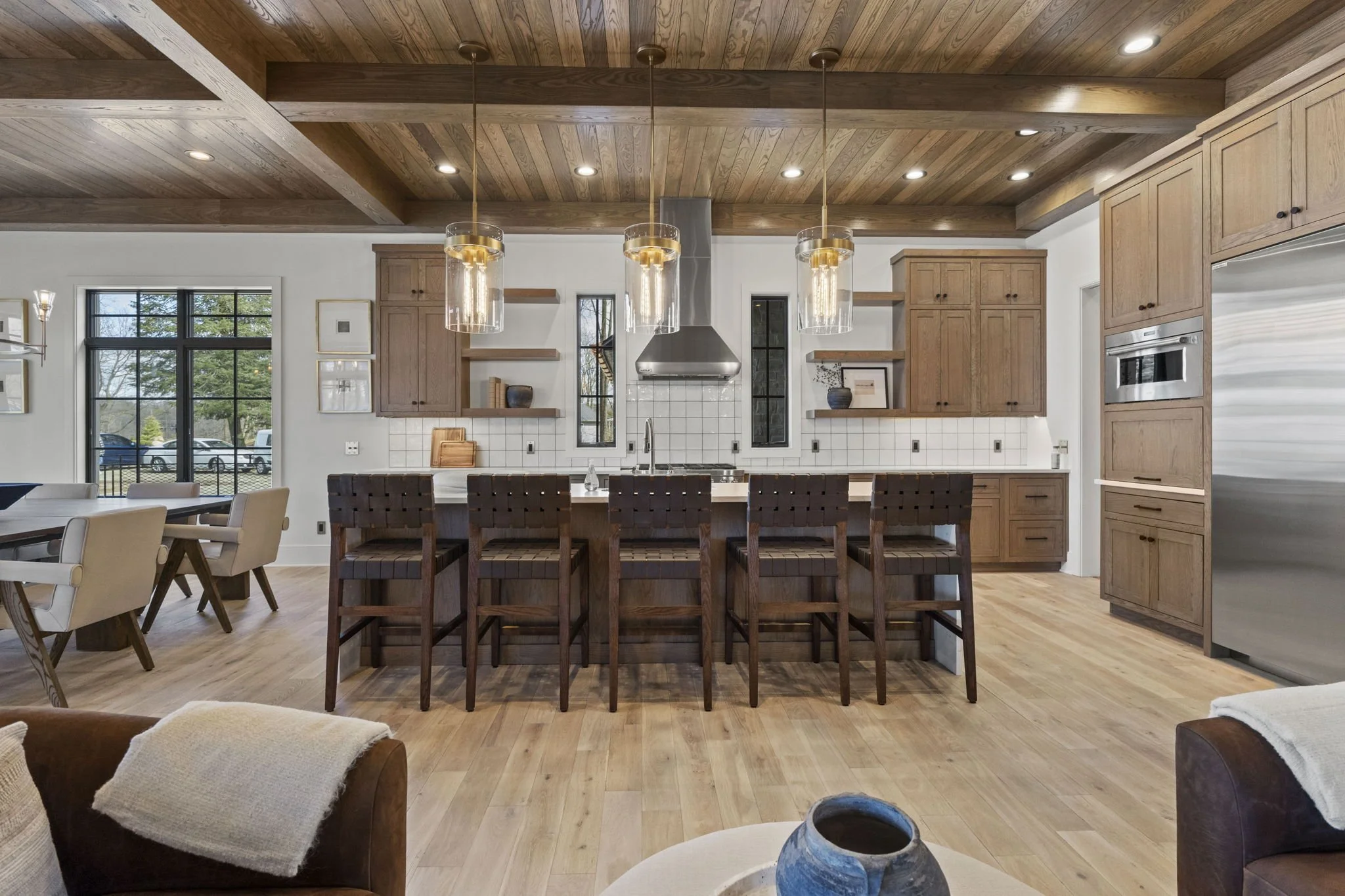 Modern kitchen with wooden cabinets, white tiled backsplash, stainless steel appliances, wooden ceiling with recessed lighting, and a kitchen island with six dark wood chairs.