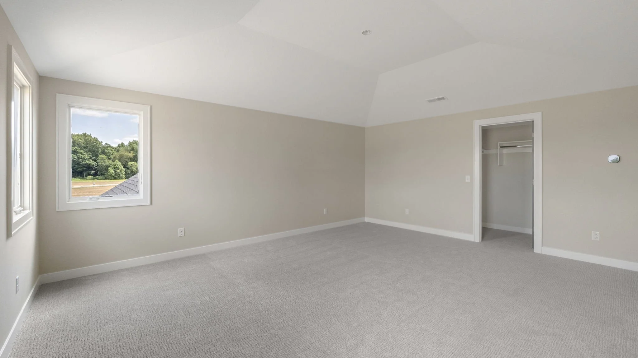 Empty bedroom with beige walls, carpeted floor, two windows with outside view of trees and sky, and an open closet door.