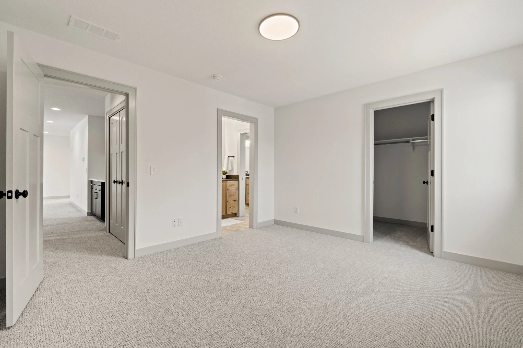 An empty bedroom with white walls, carpeted floor, open closet, and multiple doors leading to other rooms, with ceiling light fixture.