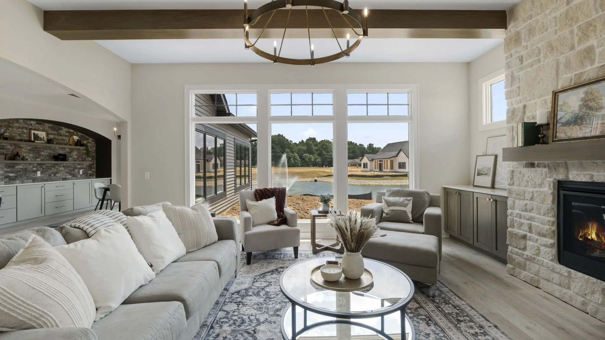 Living room with large window view of a pond, fountain, and houses outside. Light-colored furniture, fireplace, and a circular chandelier.