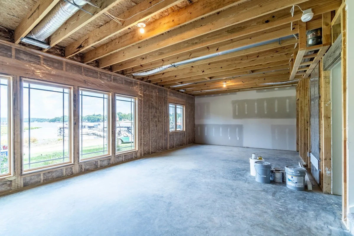 Interior of a room under construction with exposed wooden ceiling beams, new drywall, and large windows showing an outside view of boats and water.