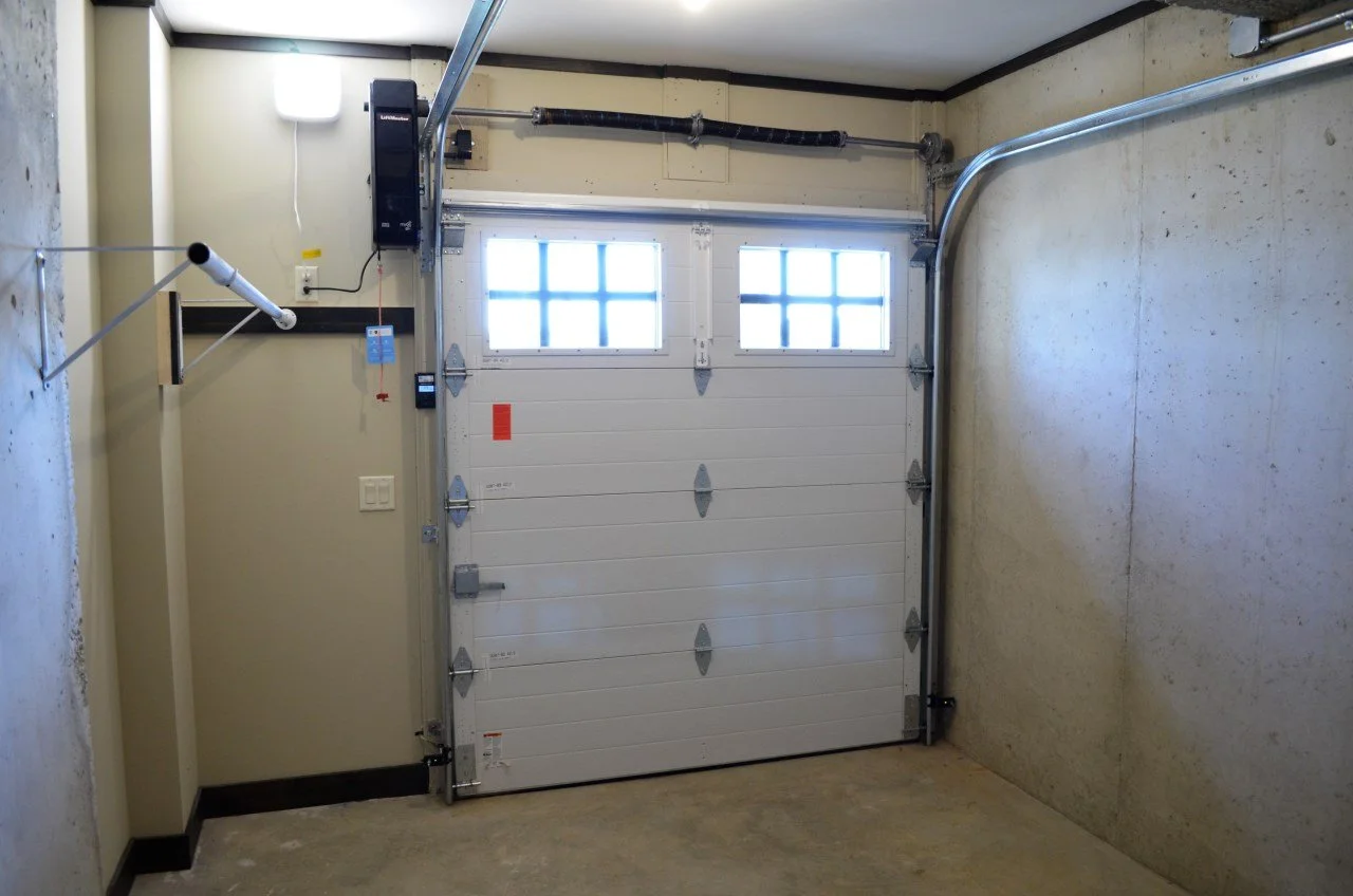Garage with a white roll-up door, small windows near the top, concrete walls, and a garage door opener mechanism.