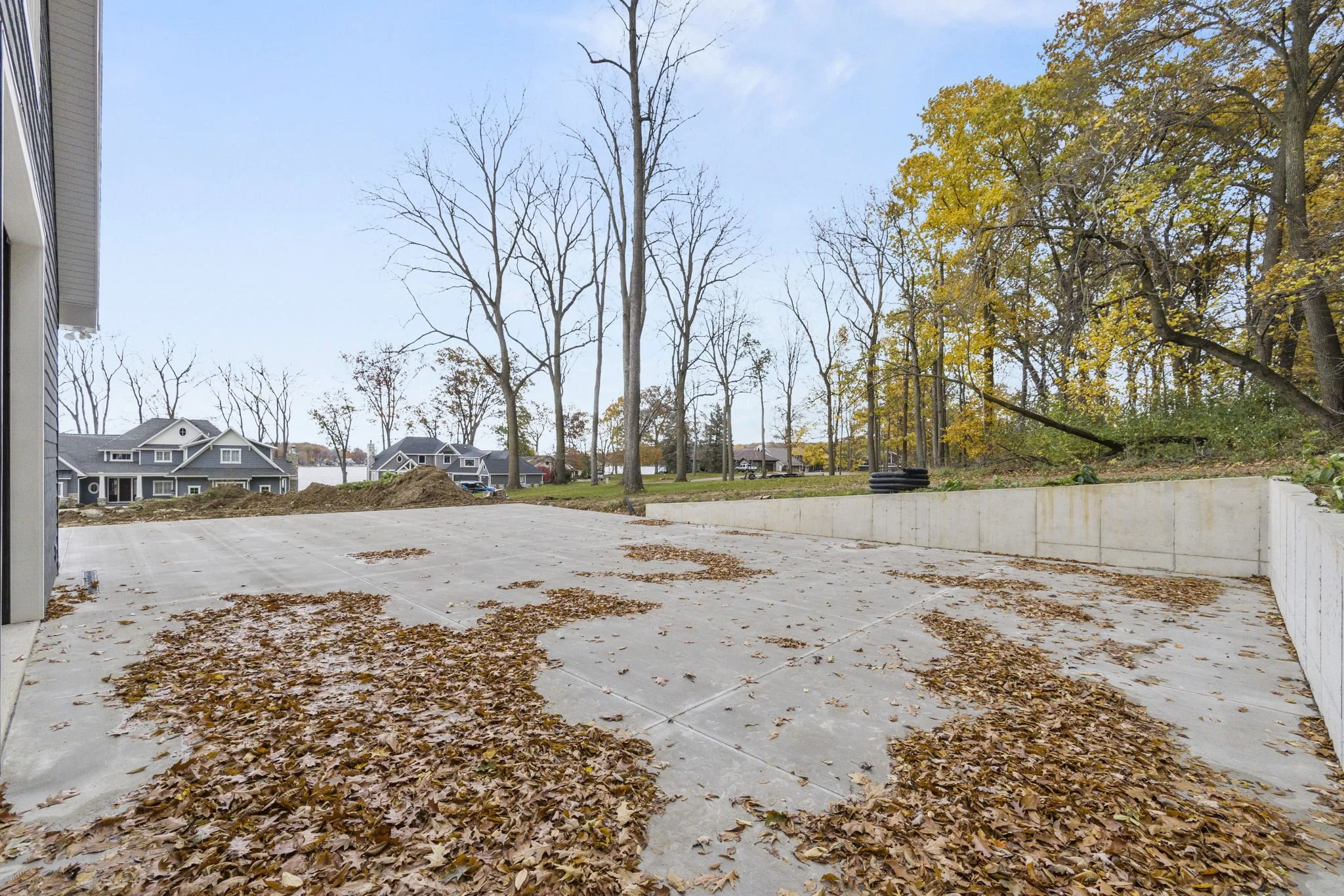 Empty concrete patio with fallen leaves, bordered by a retaining wall, with leafless trees and some with yellow leaves, in a suburban neighborhood during fall.
