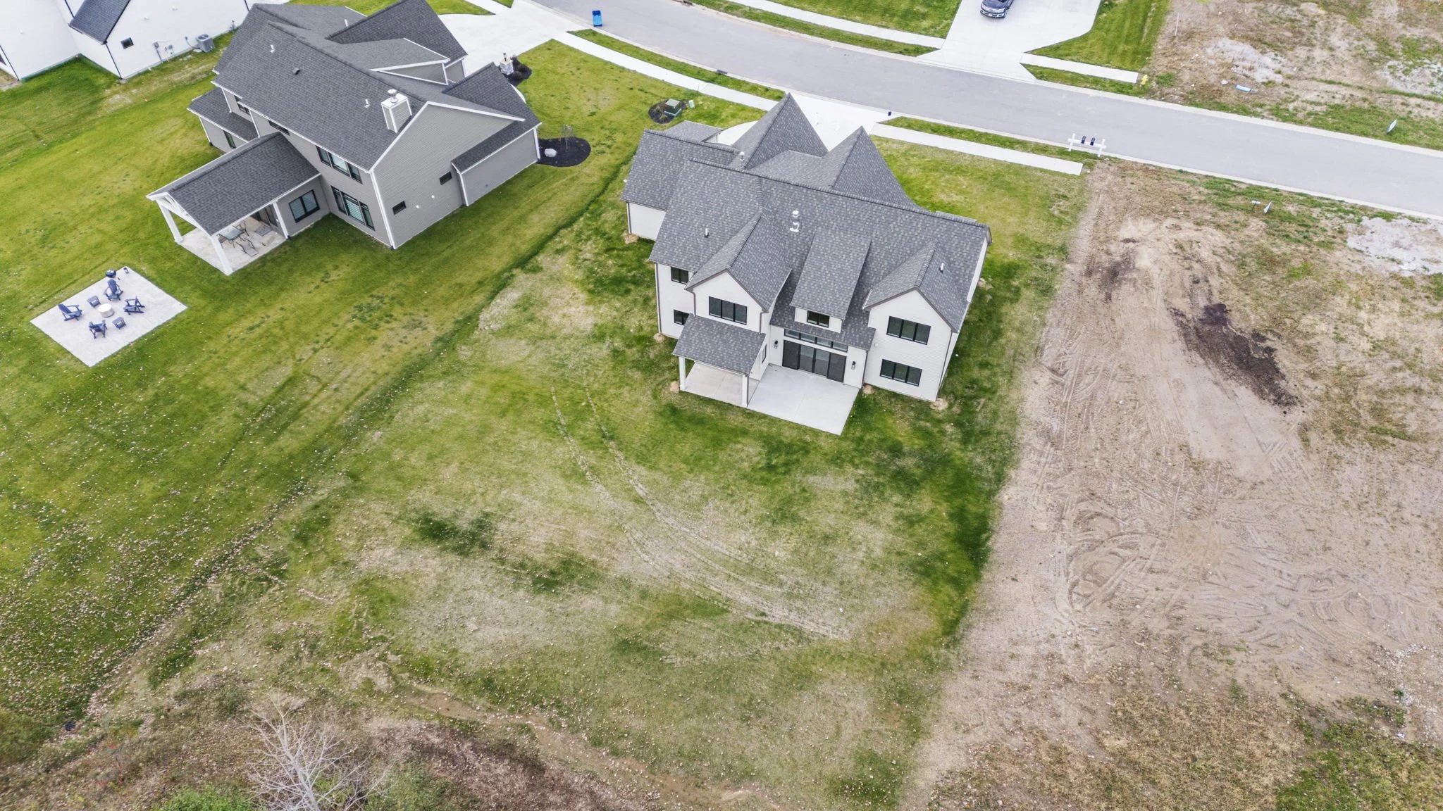 Aerial view of two modern residential houses with yards, situated in a suburban neighborhood. One house has a dark roof and the other has a light-colored exterior with a patio. The area around the houses includes grassy lawns, a paved road, and patch