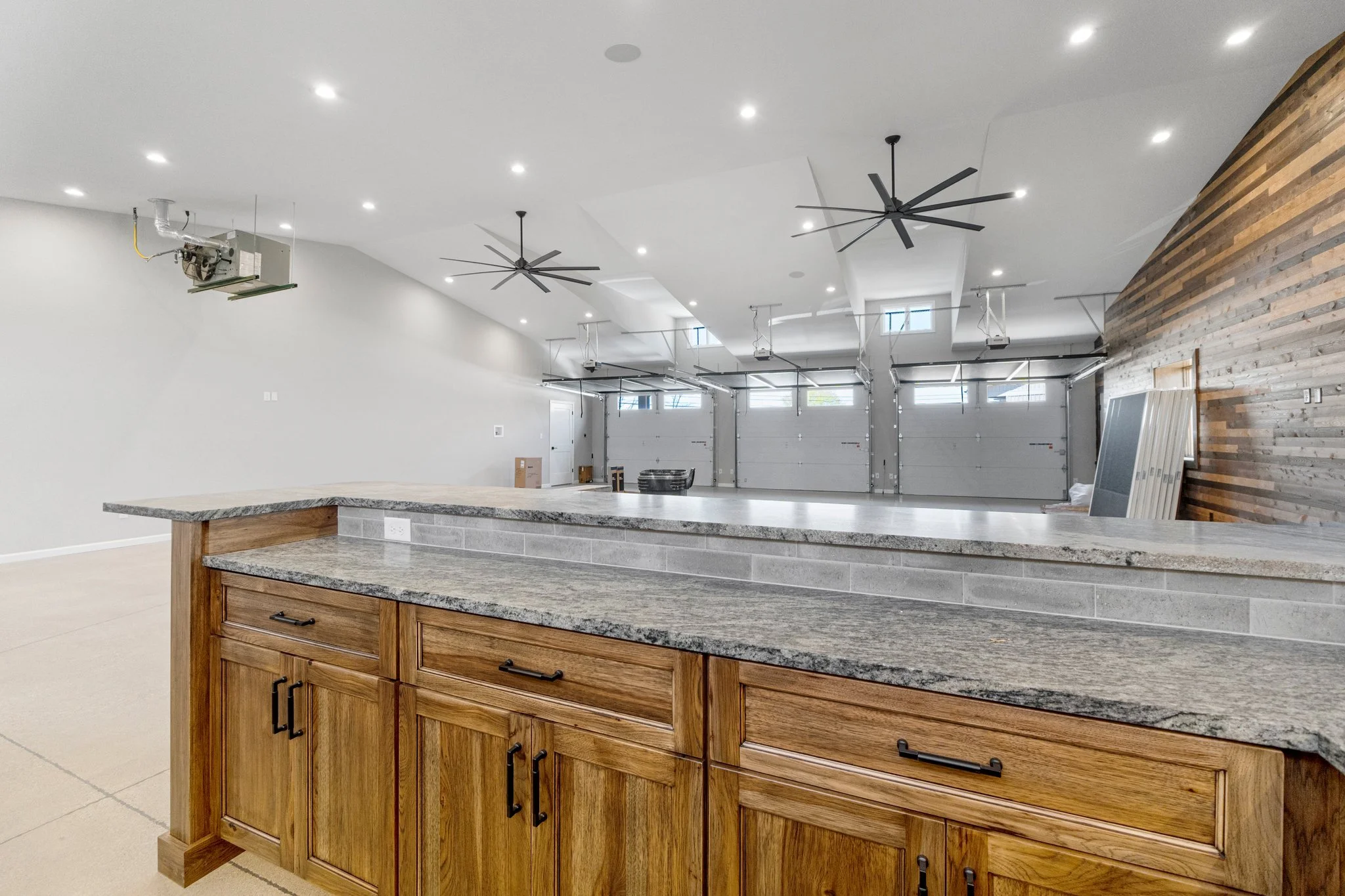 Empty modern garage with wooden kitchen island and granite countertop in the foreground, white walls, ceiling fans, recessed lighting, garage doors, and wood accent wall.