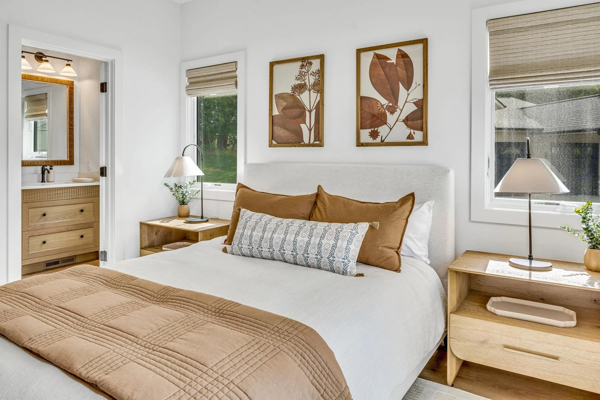 Cozy bedroom with a white bed adorned with brown and patterned pillows, wooden nightstands, framed botanical artwork, side lamps, and windows with beige Roman shades.