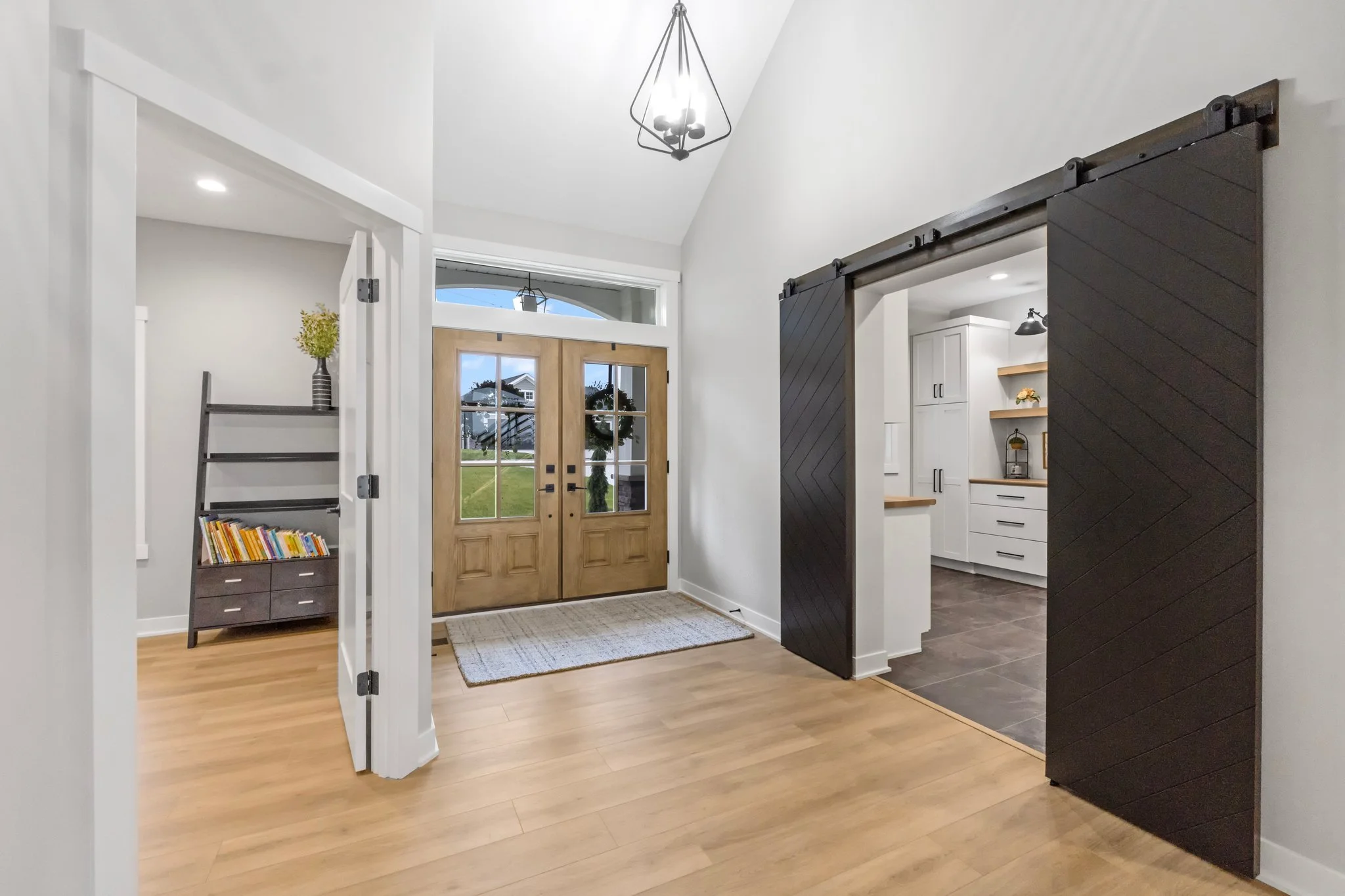 Entryway with wooden double doors with glass panels, light wood floors, and modern decor. A black metal chandelier hangs from the ceiling. To the left, a small bookshelf with children’s books, and to the right, a dark sliding barn door reveals the ki