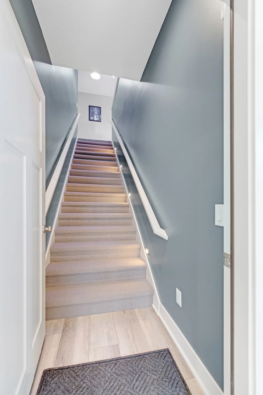 Interior view of a staircase with beige carpet, blue walls, white trim, and recessed lighting, leading to an upper floor with a framed picture on the wall.