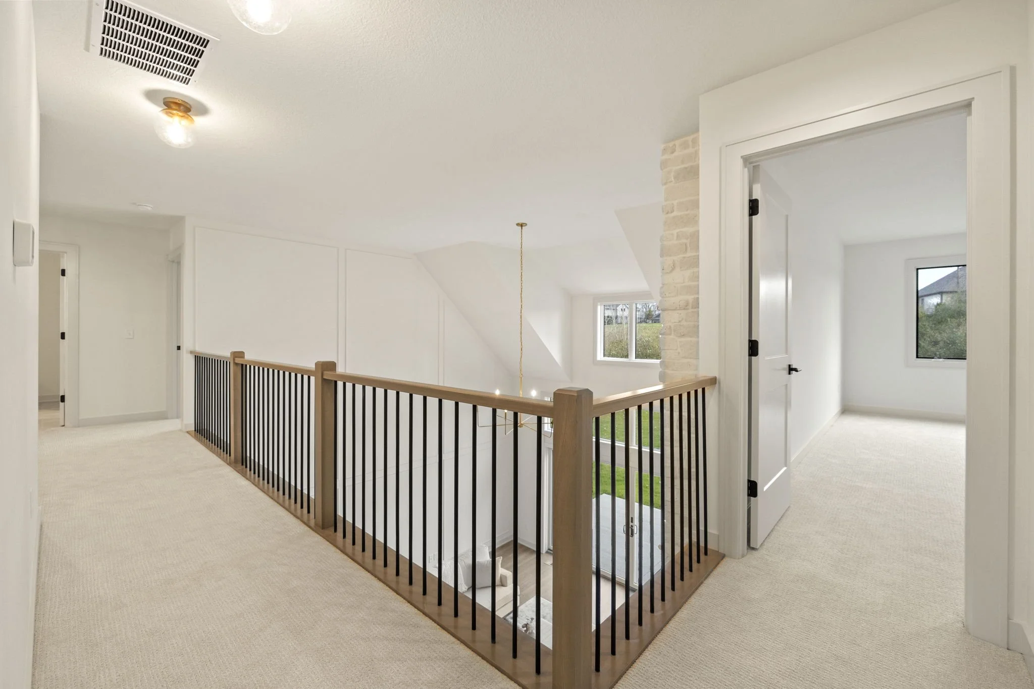 Bright, white upper-level hallway with beige carpet, black and wood railing around a staircase, and three open doors leading to bedrooms with large windows.