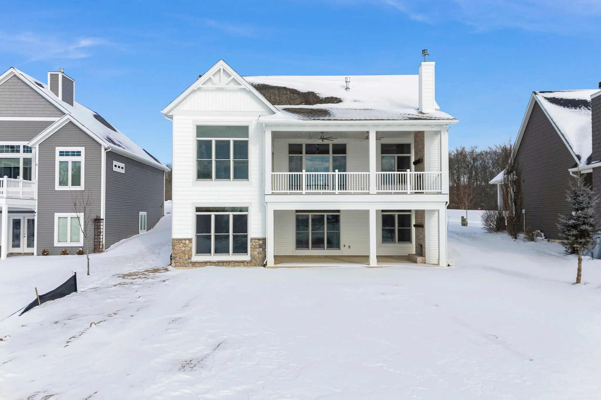 A white, three-story house with large windows, a covered back porch, and a snowy yard, flanked by similar gray houses under a blue sky.
