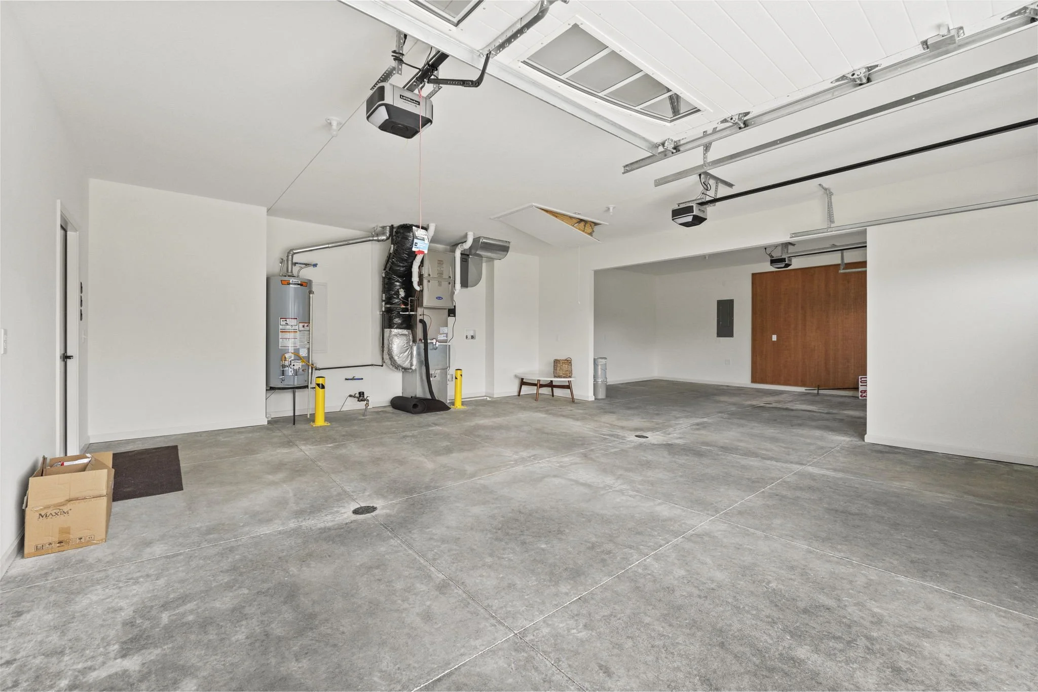 Empty garage with concrete floor, white walls, and attached garage door. There are pipes, a water heater, and HVAC ducts on the left wall, and two garage door openers on the ceiling.