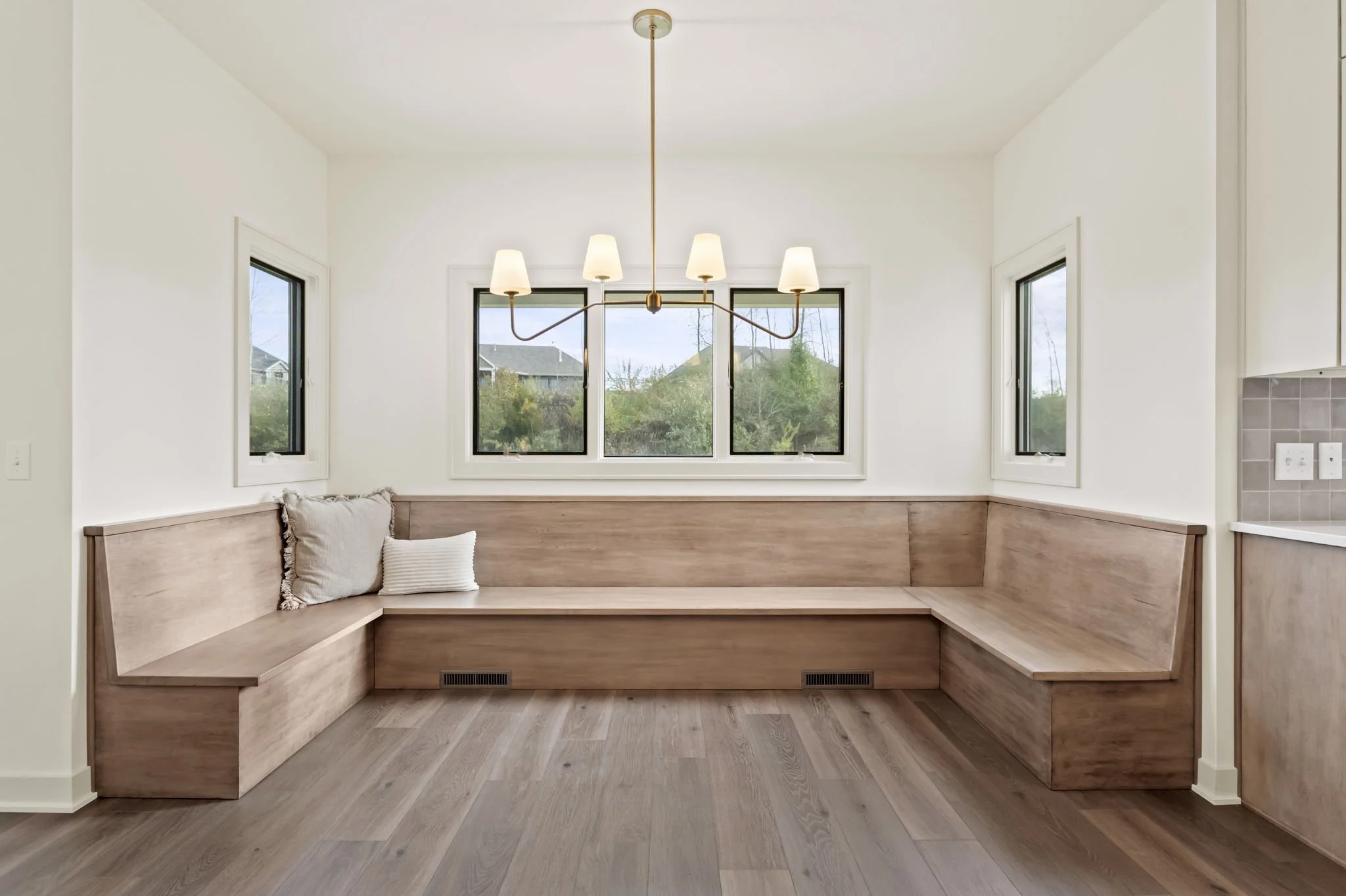 A cozy kitchen nook with built-in wooden bench seating, two throw pillows, three windows, and a modern chandelier with five shades overhead.