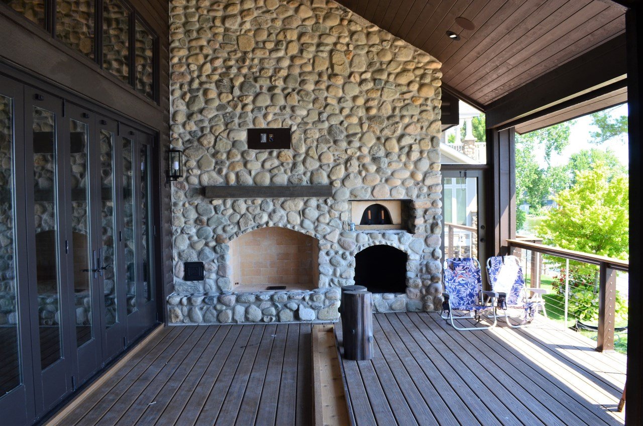 Outdoor porch or patio with a stone fireplace, rocking chairs, and a view of green trees.