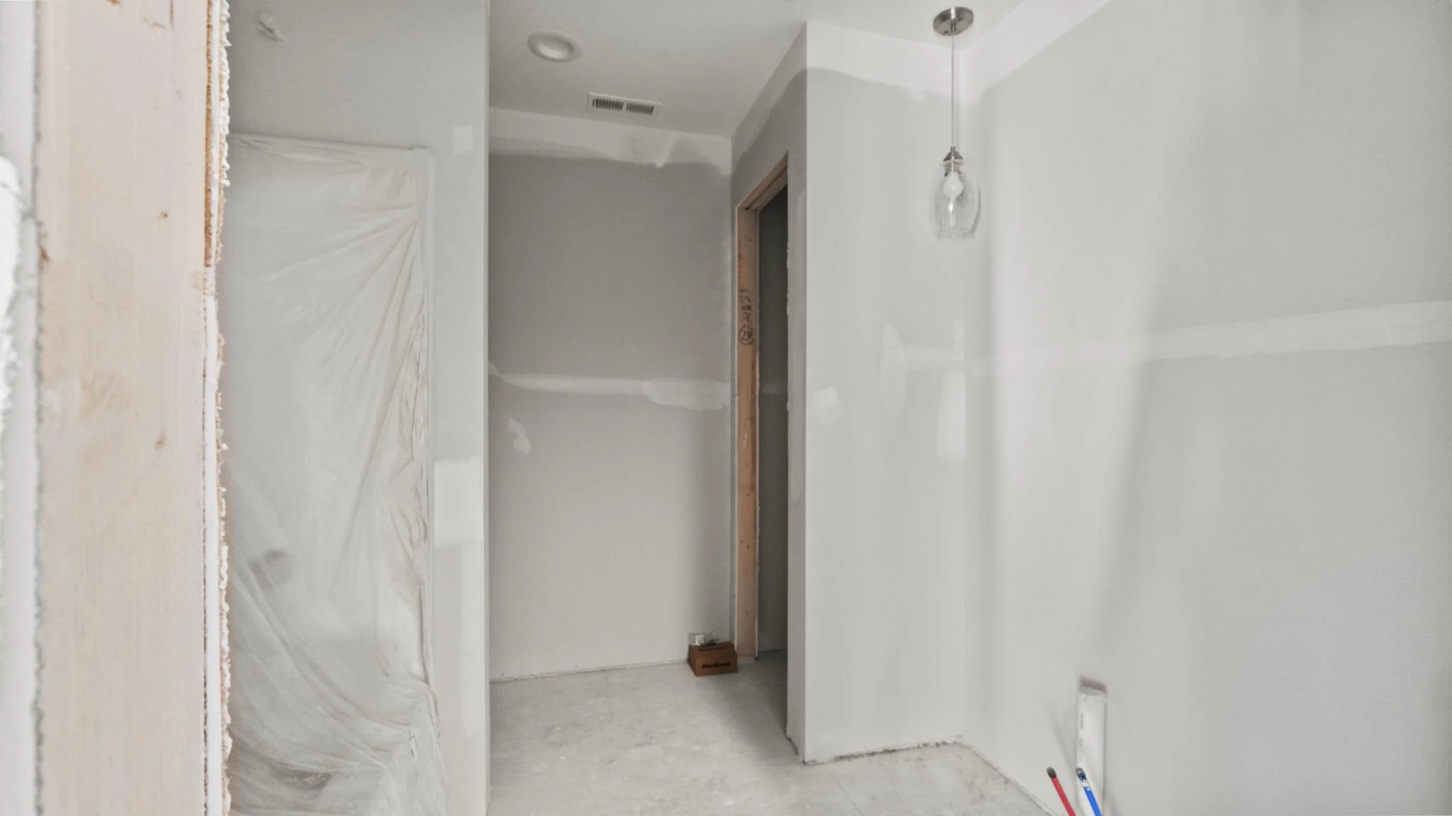 Interior of a room under construction with white drywall, an open doorway with exposed wood frame, protected plastic sheet on a left wall, a hanging light fixture, and construction tools on the floor.