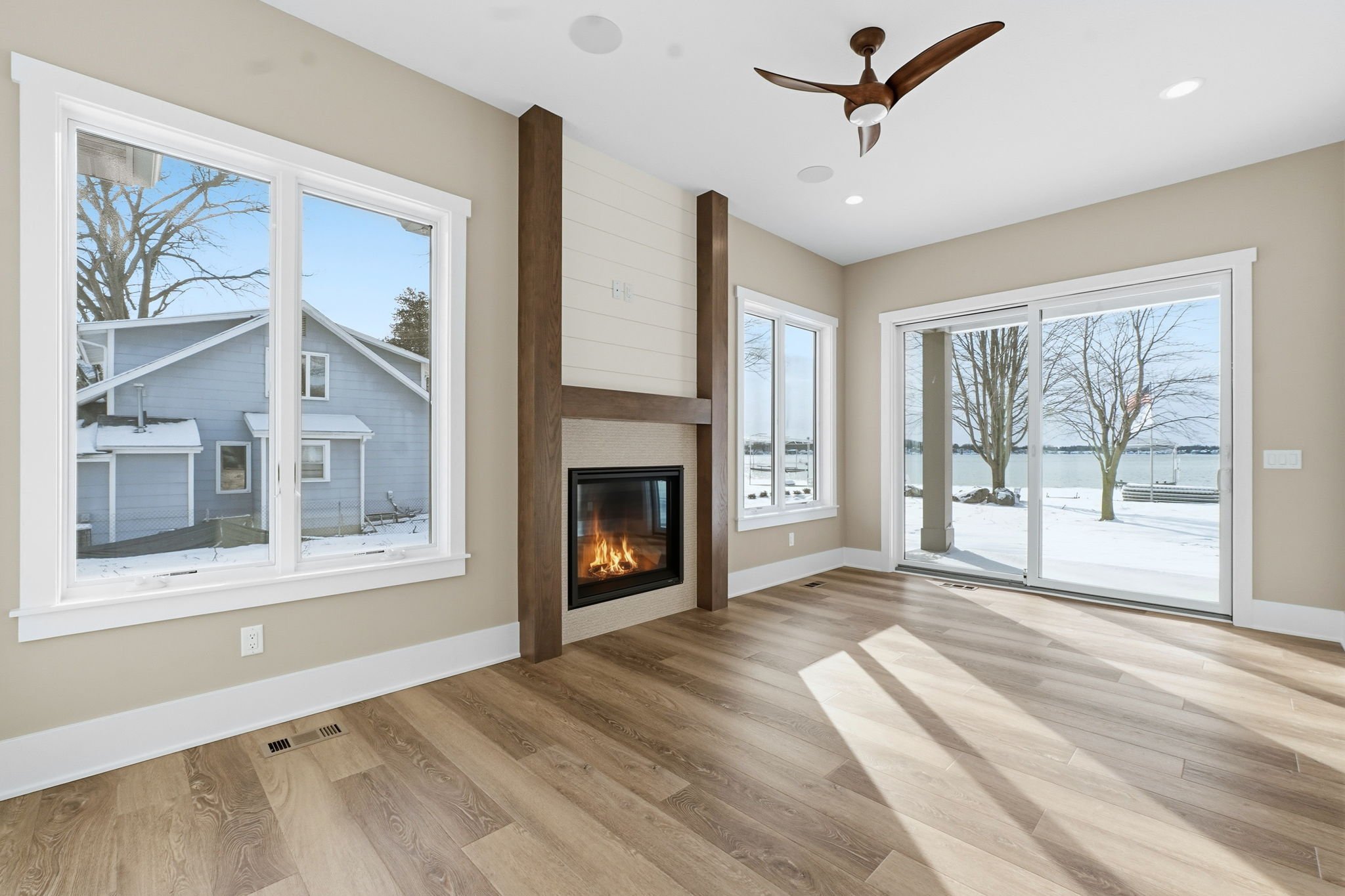 Empty living room with large windows, sliding glass door, fireplace, beige walls, wooden floors, and ceiling fan, overlooking a snowy landscape.
