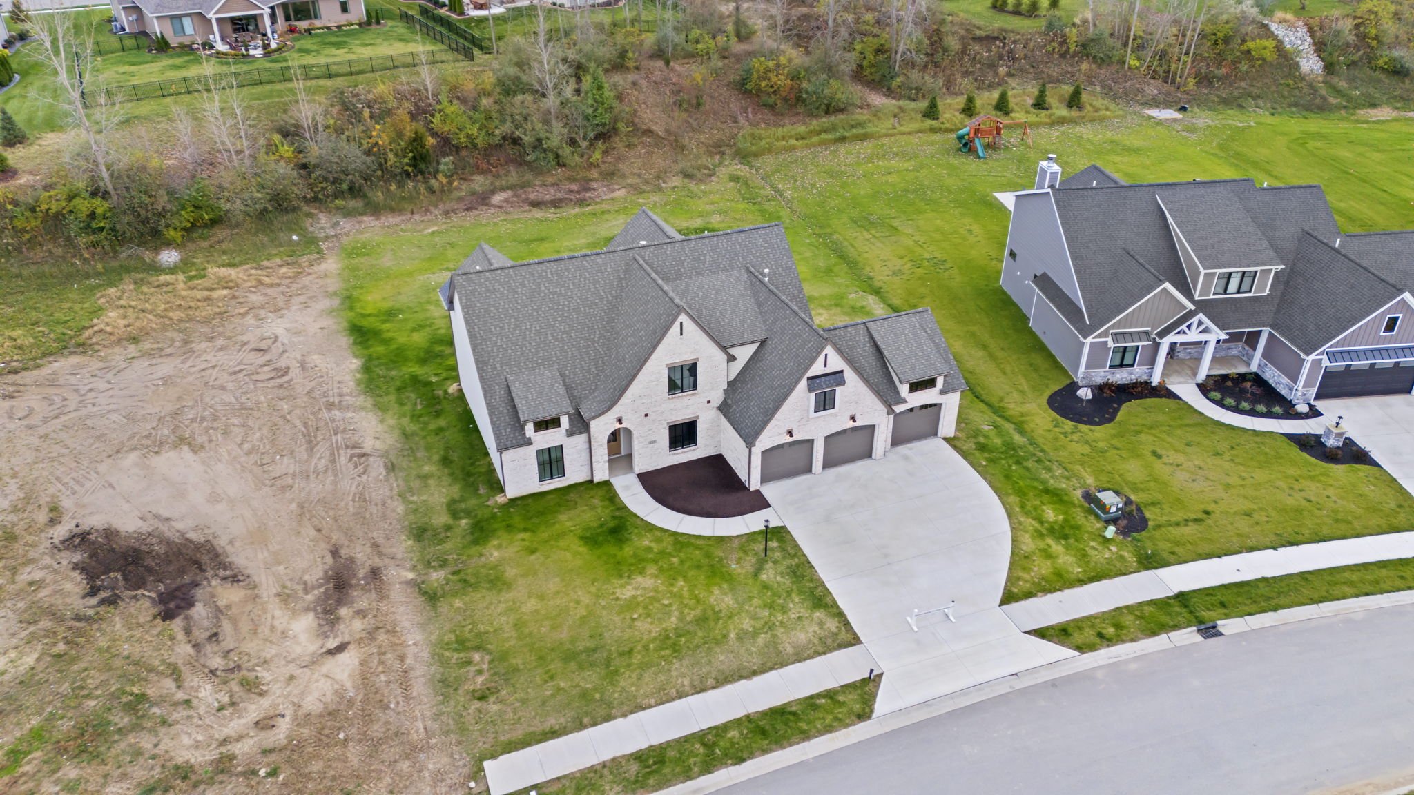 A newly built two-story house with a gray roof, white brick exterior, and a concrete driveway. The house has a curved front walkway, a small front yard, and neighboring houses nearby. There is an empty dirt lot on the left side and a playground in th