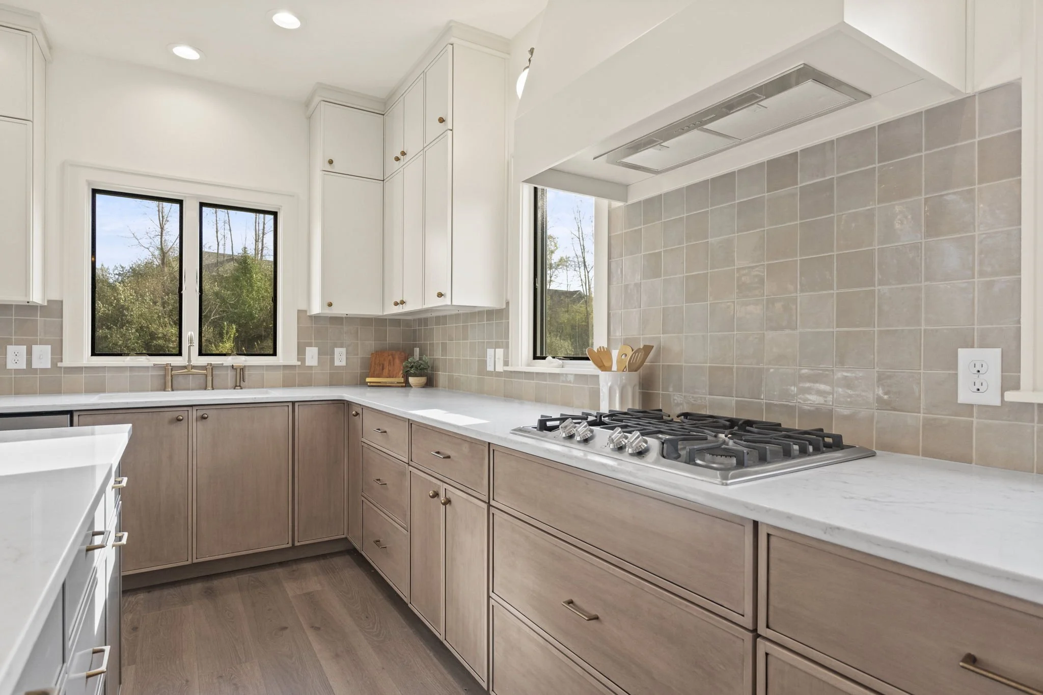 A modern kitchen with white upper cabinets, beige lower cabinets, and a gas cooktop on a white marble countertop. There are two windows above the sink and a beige tiled backsplash.