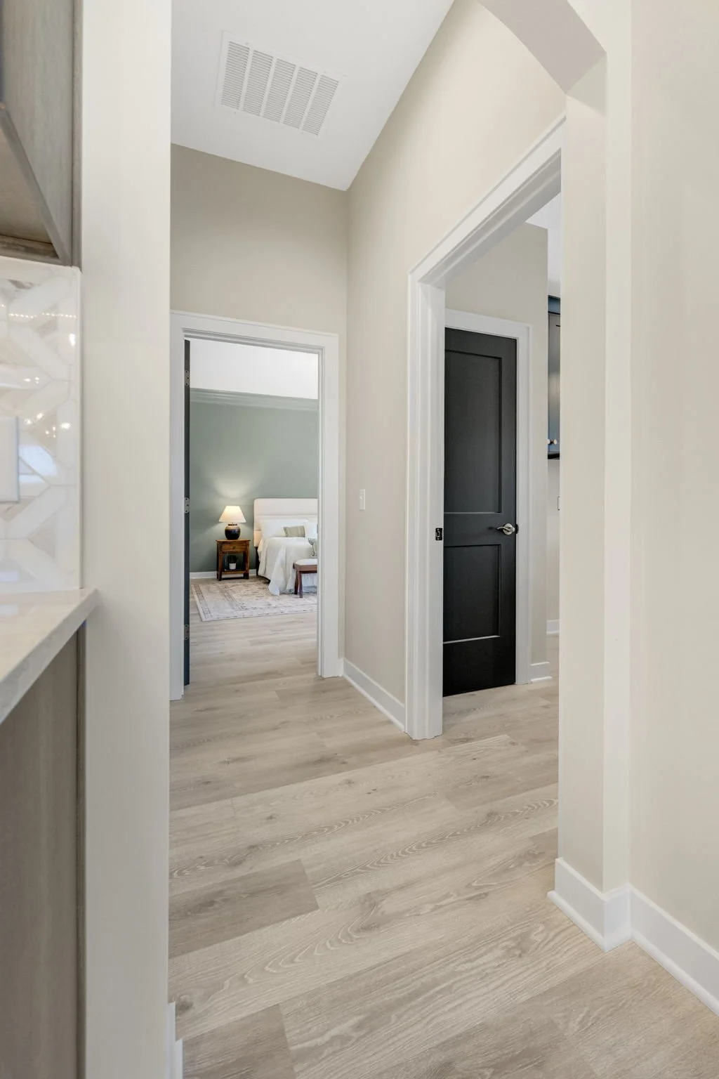 View of a hallway leading to a bedroom with a nightstand, lamp, and bed, featuring beige walls and wooden flooring.
