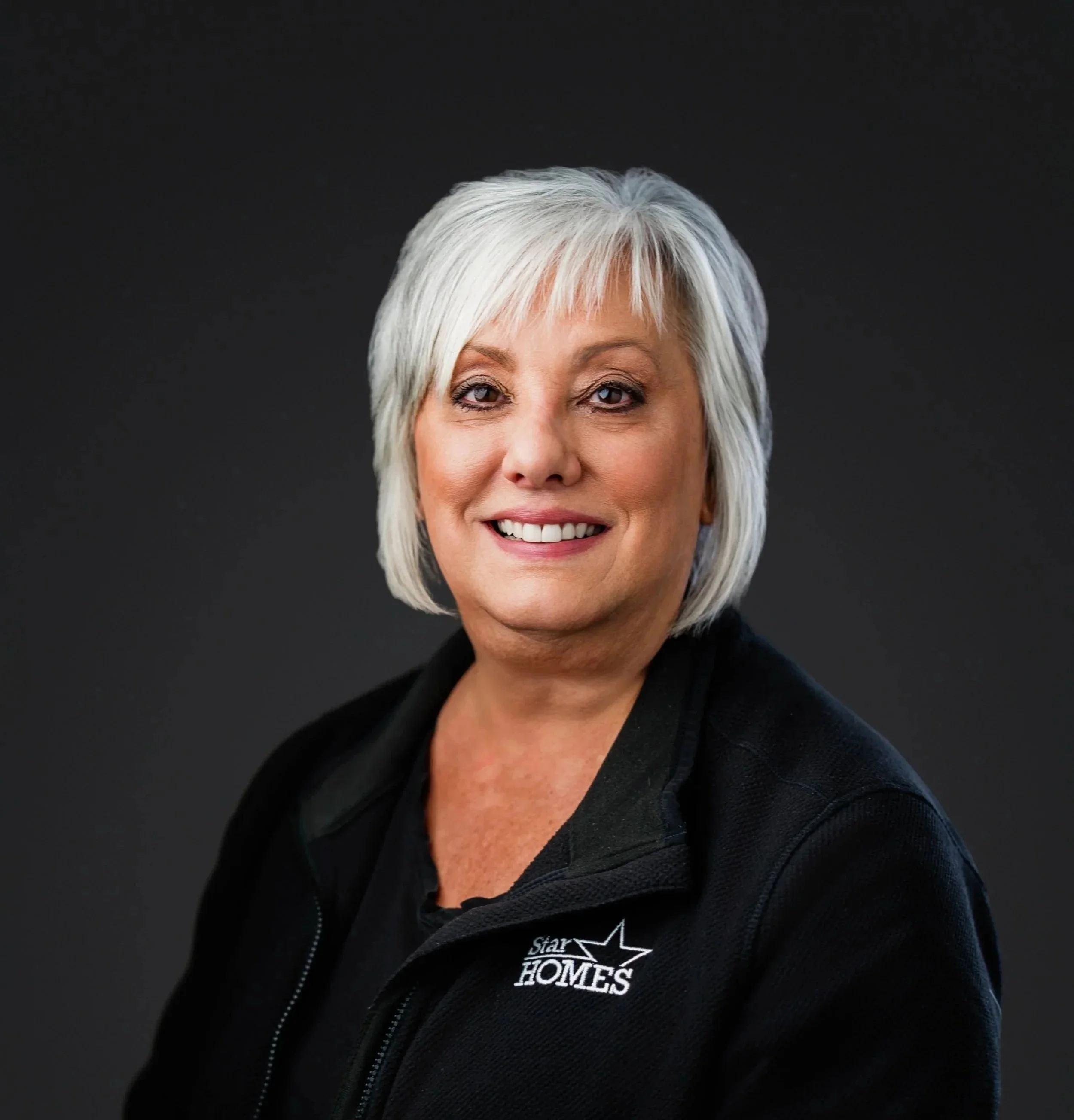 A woman with short, silver hair and a friendly smile is wearing a black zip-up jacket with a 'Star HOMES' logo on it. The background is plain black.