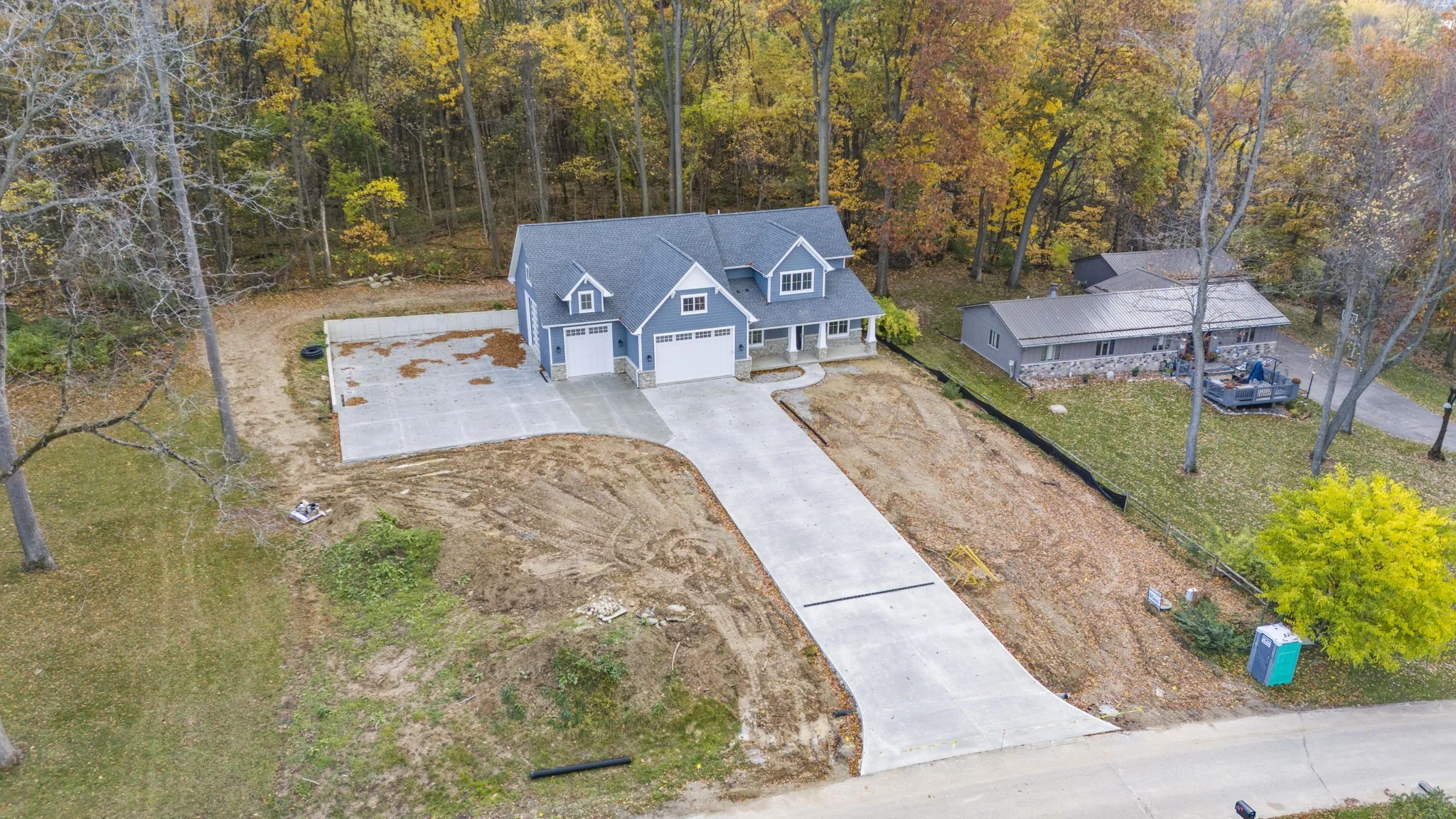A newly constructed blue house with a three-car garage, surrounded by a spacious yard under construction, with nearby wooded area.