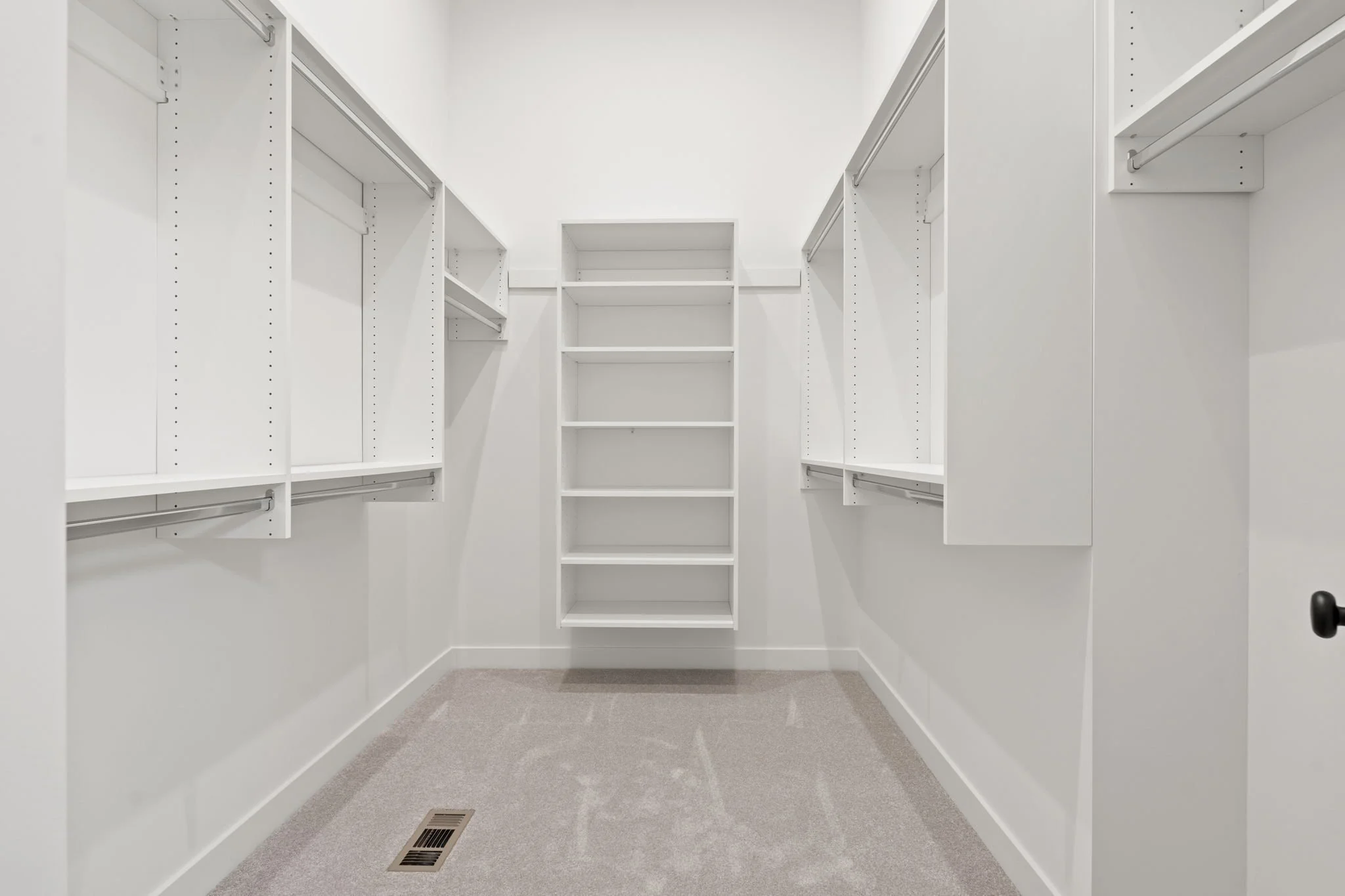 Empty walk-in closet with white shelves and hanging rods on both sides, beige carpeted floor, and a central bookshelf with open shelves.