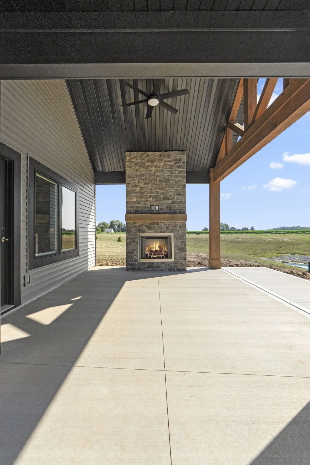 Covered outdoor patio with a stone fireplace, ceiling fan, and wooden beams, overlooking a grassy field under a blue sky.