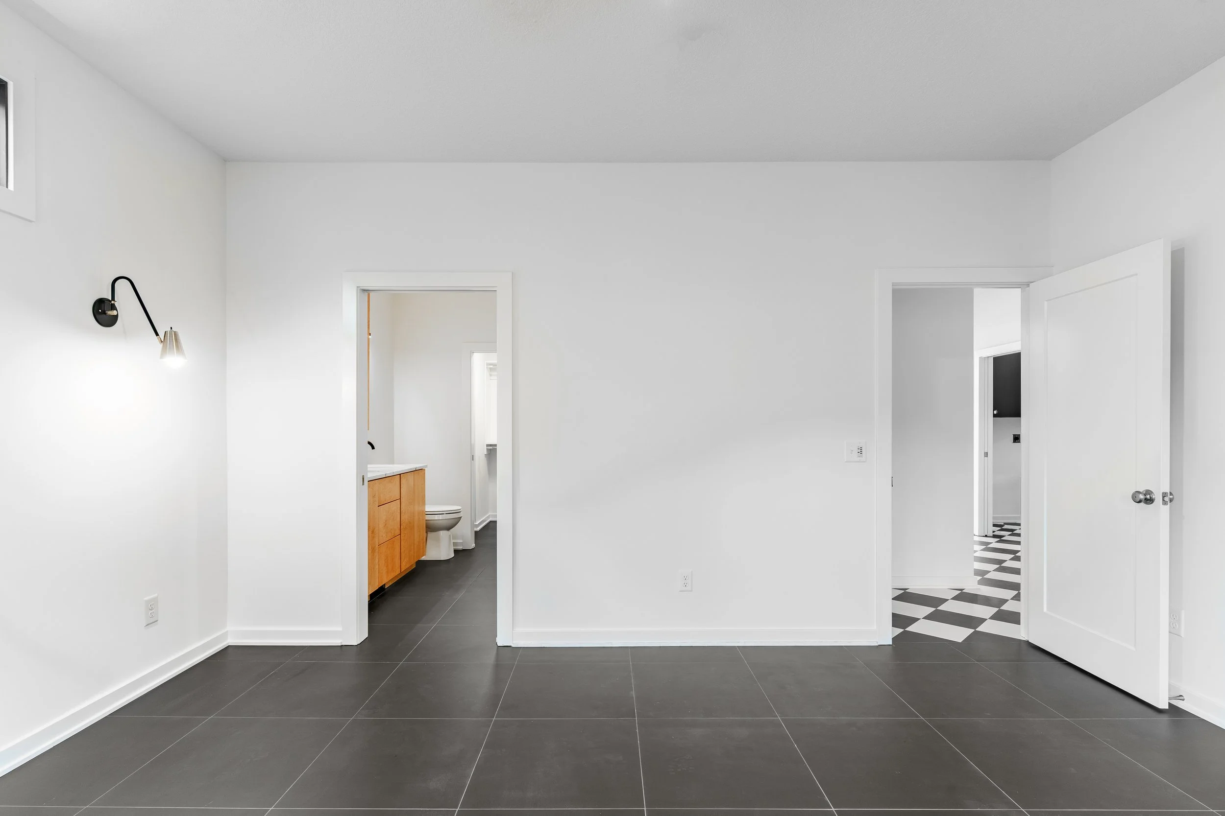 Empty room with black tile flooring, white walls, and two open doorways leading to a bathroom and a hallway. The bathroom has a wooden vanity and a toilet, with a small window above.