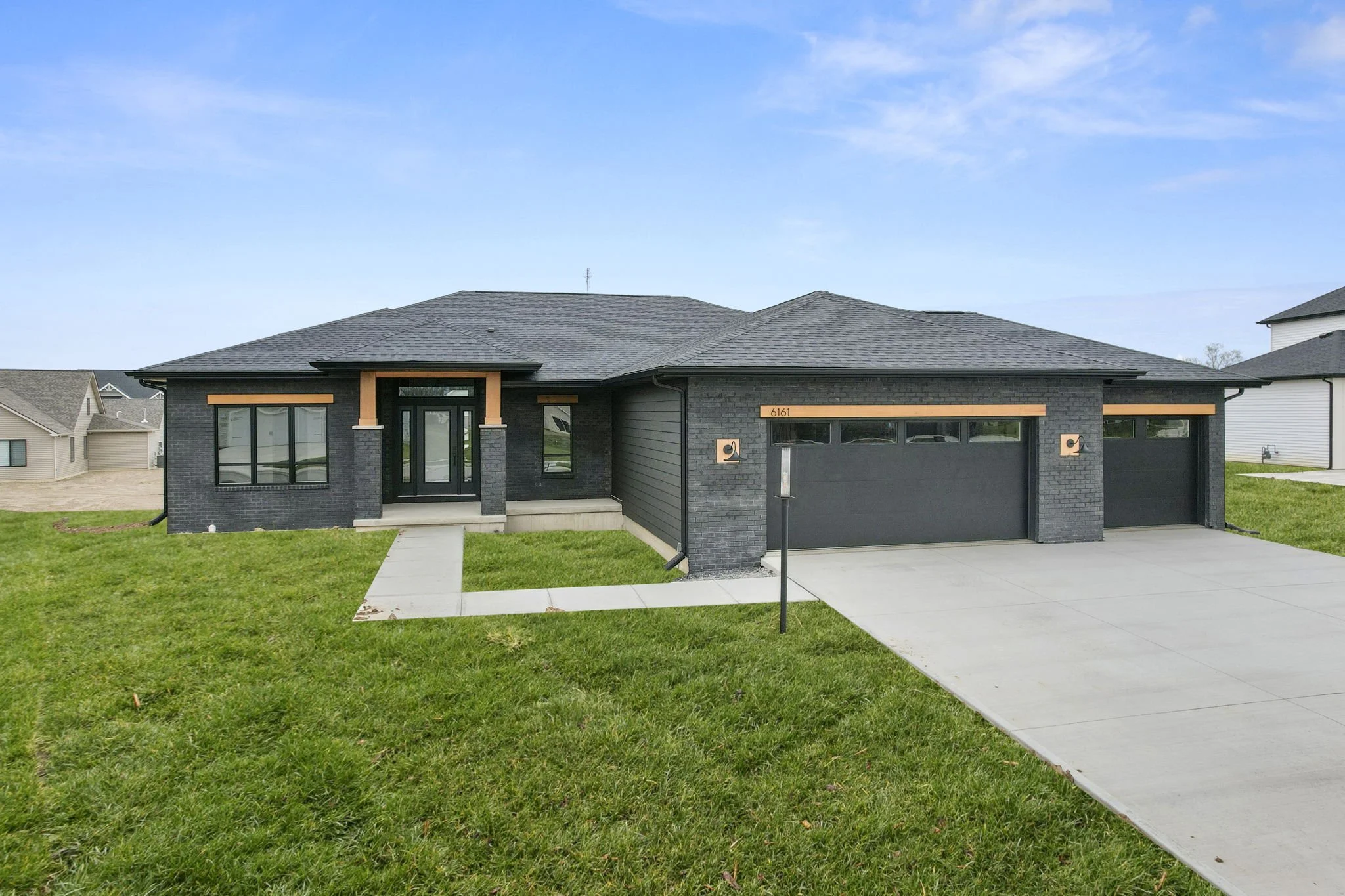 A modern single-story house with a black brick exterior, gray roof, and a two-car garage. The house has a small front porch with concrete steps, a green lawn, and a sidewalk leading to the front door.