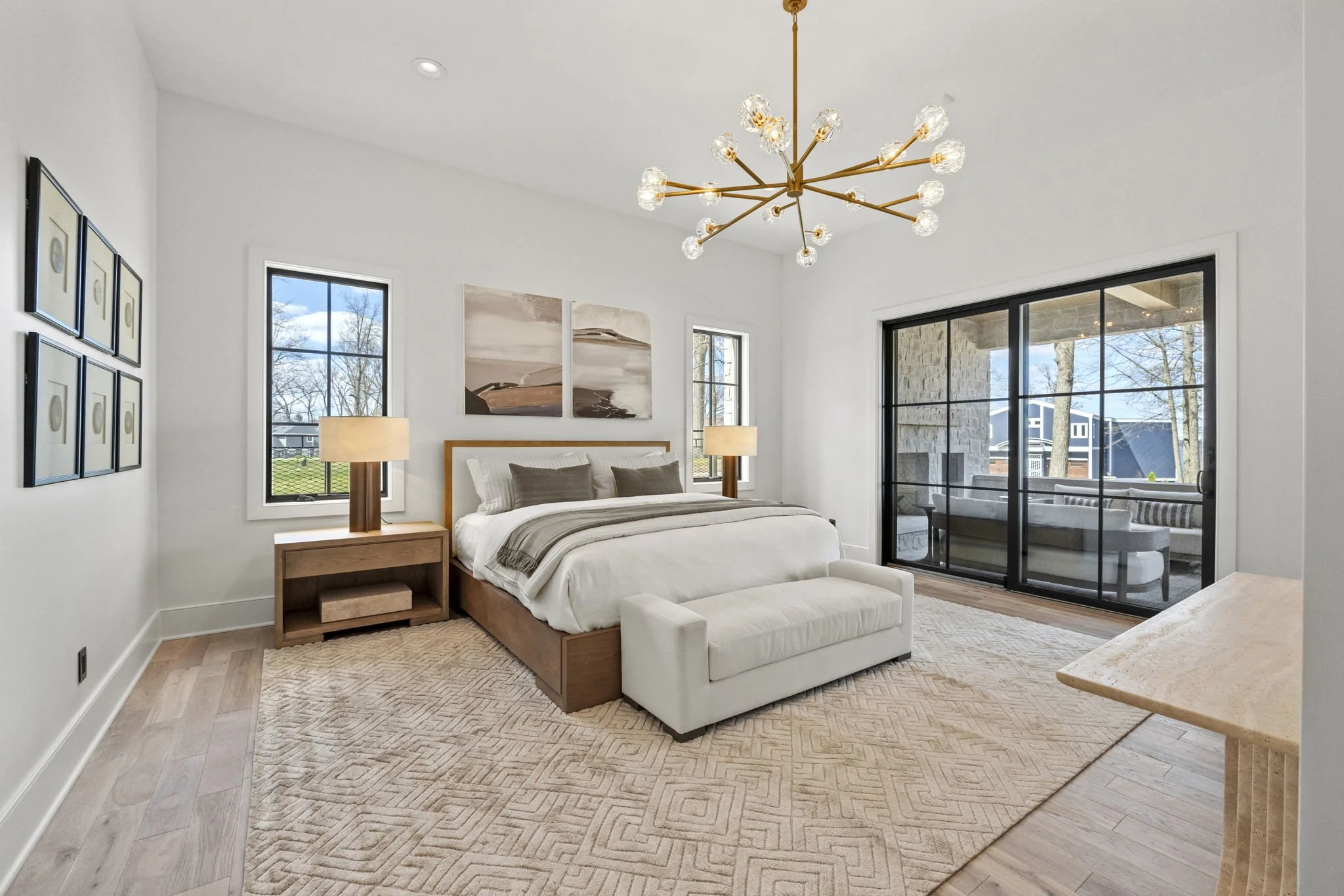 A modern bedroom with a large bed, two nightstands with lamps, a white loveseat at the foot of the bed, art on the walls, a chandelier, and a sliding glass door leading to a balcony with outdoor seating.