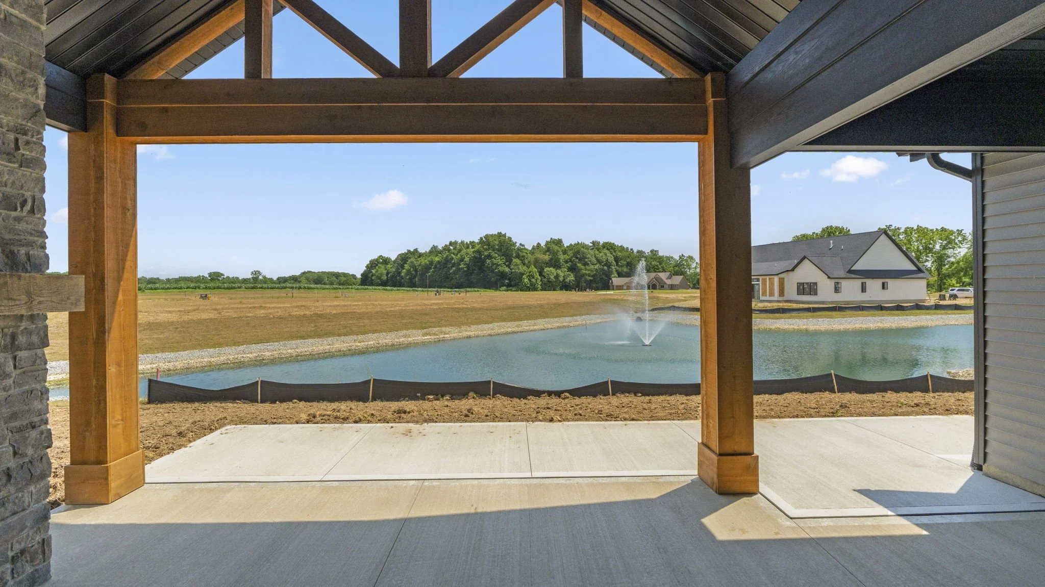 View from a covered patio showing a pond with a fountain, a grassy field, and a partially constructed house in the background under a bright blue sky.