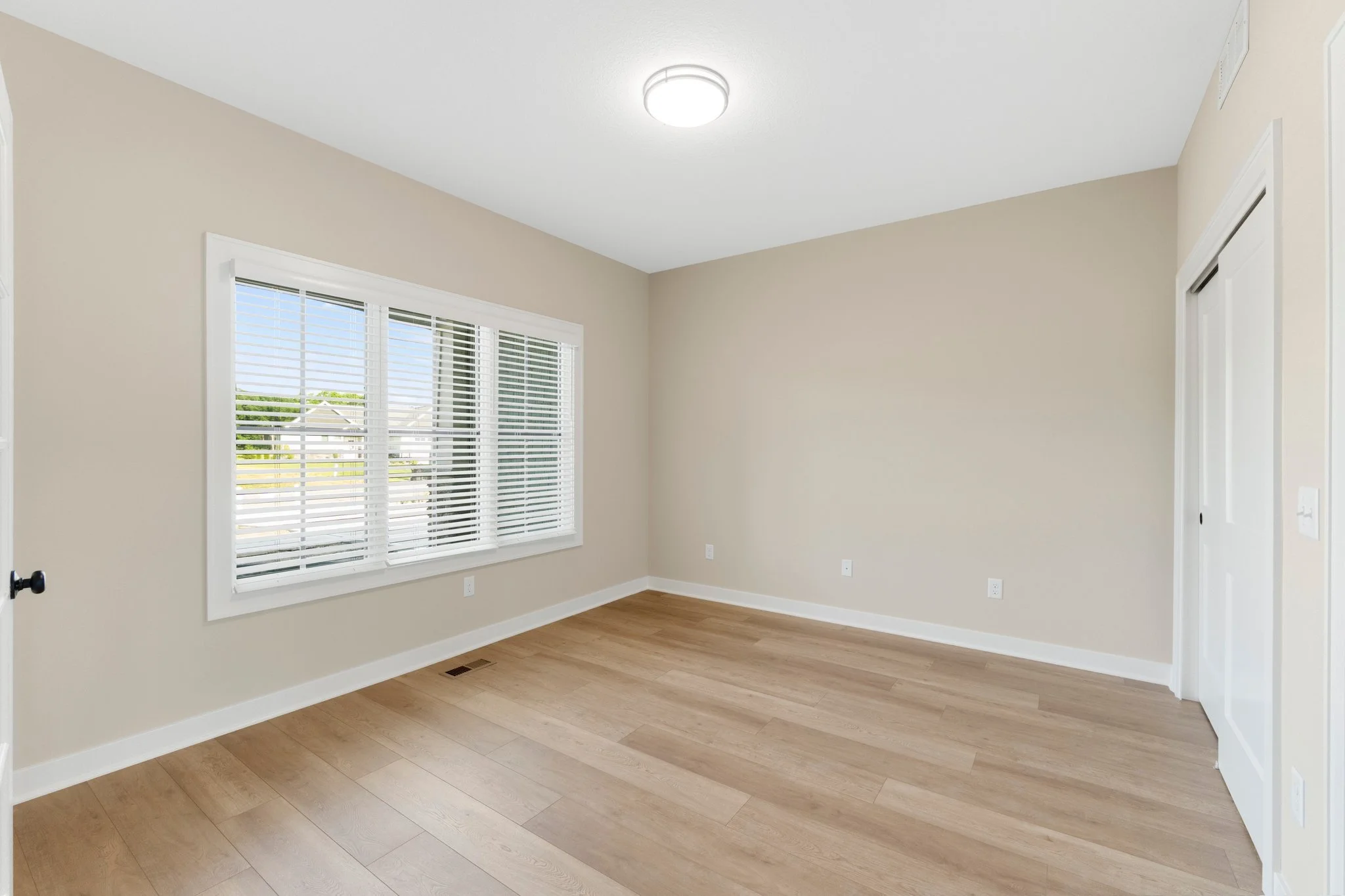 Empty room with large window and beige walls, hardwood floor, and white baseboards and trim.