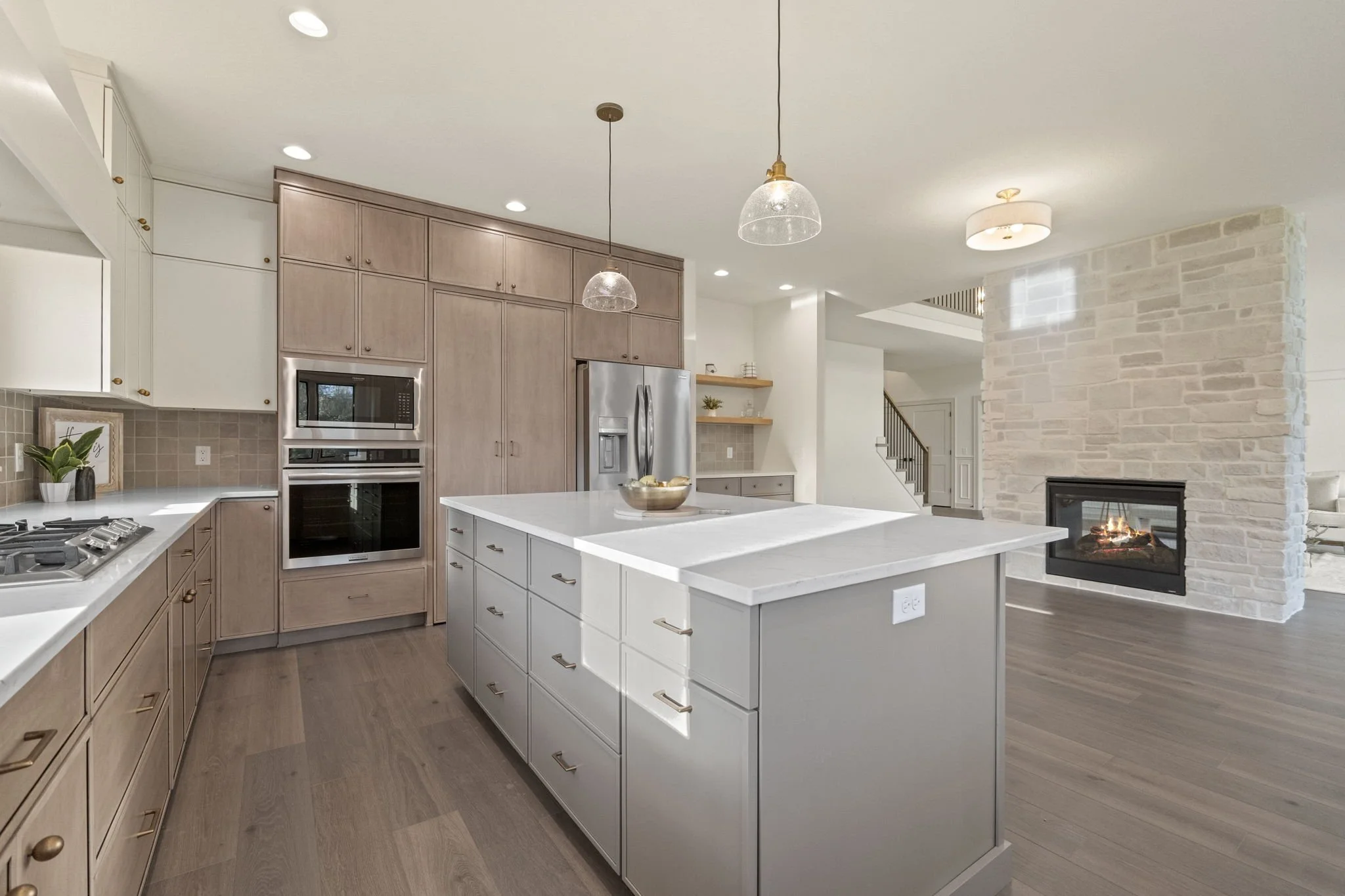 Modern kitchen with a large white island, light wood cabinets, stainless steel refrigerator, built-in oven and microwave, and a fireplace in the adjacent living area.