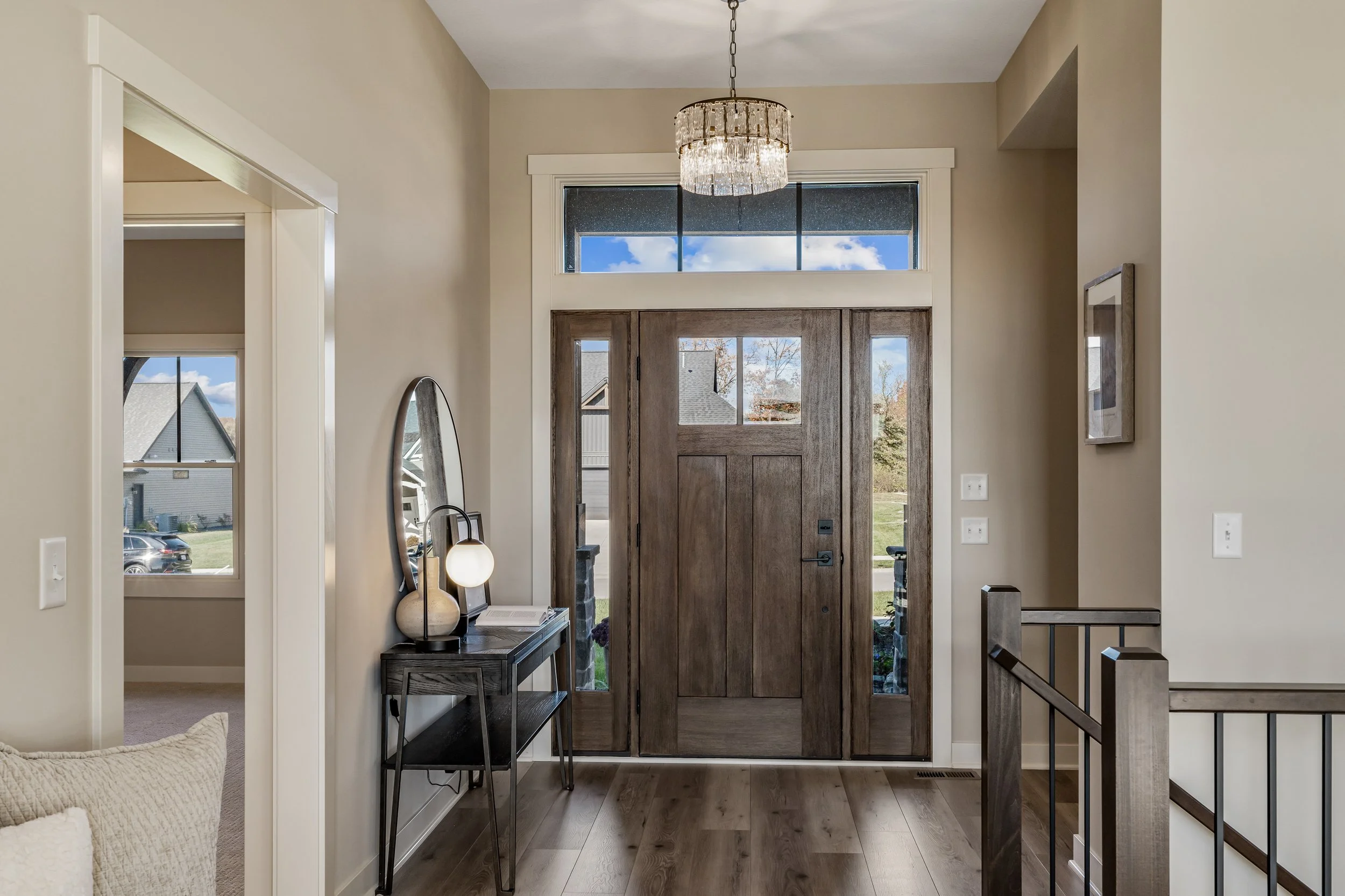 Entryway with a wooden front door, a transom window above, and sidelights, a chandelier hanging from the ceiling, a dark console table with a mirror and decorative lamp, wood flooring, and a staircase with a wooden handrail.